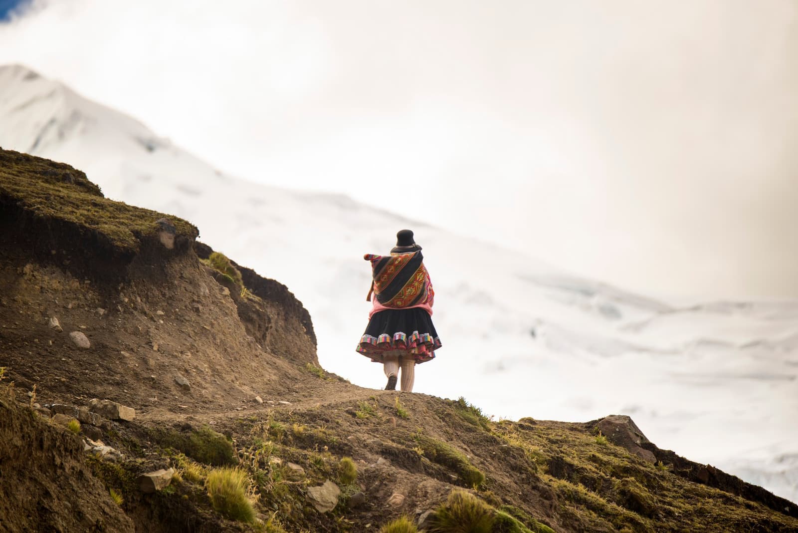 She knows this mountain the way most of us know our own street. A woman in traditional Andean dress — a colorful woven shawl, black skirt with embroidered trim, and bowler hat — walks along a high-altitude dirt trail on the Ausangate Lodge Trek, with a snow-covered peak rising behind her