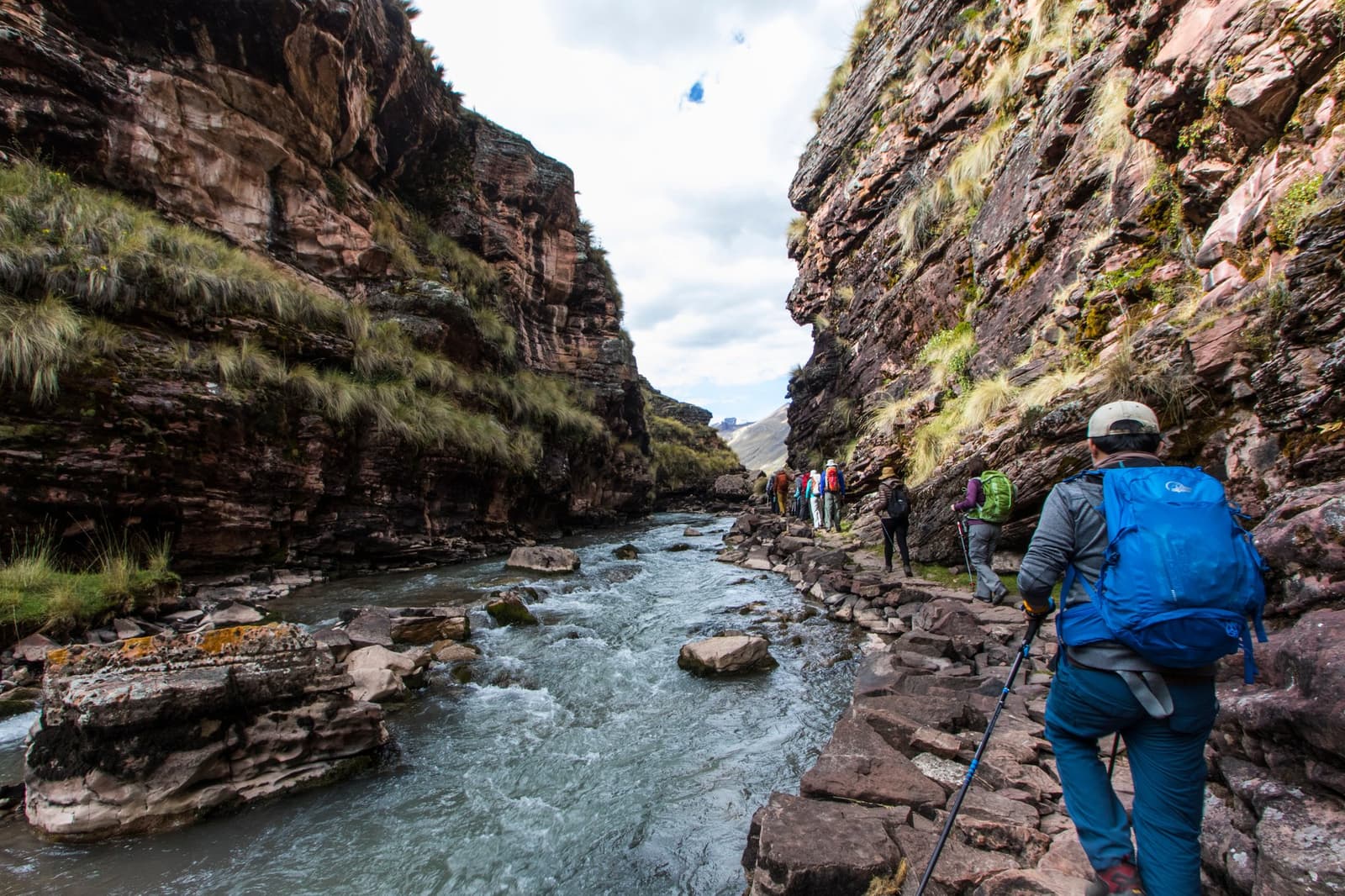 The canyon keeps you honest, single file, eyes on the rocks. Photo: Diego Nishiyama - Fotografia A group of trekkers with trekking poles and large backpacks pick their way along a rocky riverbank inside a narrow Andean canyon on the Ausangate Lodge Trek, with a fast-flowing glacial river rushing between towering stratified rock walls patched with highland grasses