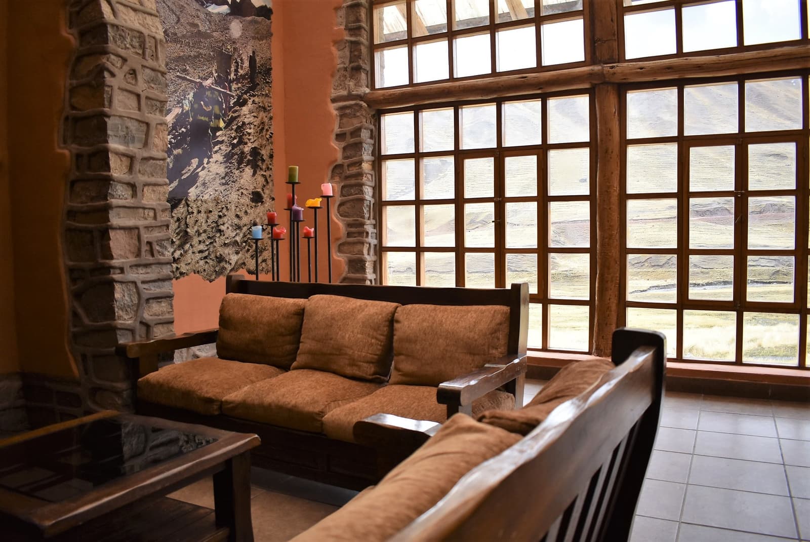Cold outside, comfortable in here. The lodge earns its keep. Photo: Warm stone-walled lounge of an Ausangate Lodge Trek mountain lodge, with wooden-framed sofas, colorful candleholders, and a tall grid window overlooking a high-altitude Andean valley