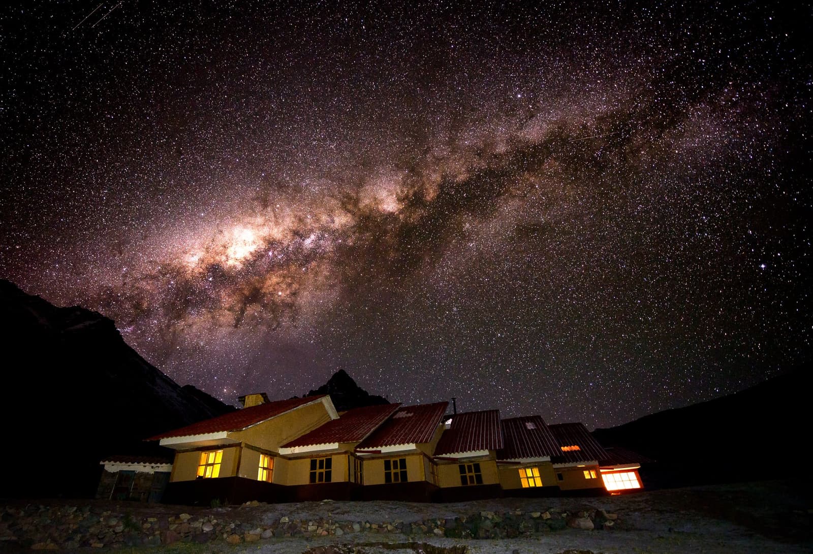 A billion stars, one warm lodge, and no light pollution for miles. The Milky Way core arching over a lit yellow-walled mountain lodge with red roofs on the Ausangate Lodge Trek, dark Andean peaks silhouetted on the horizon