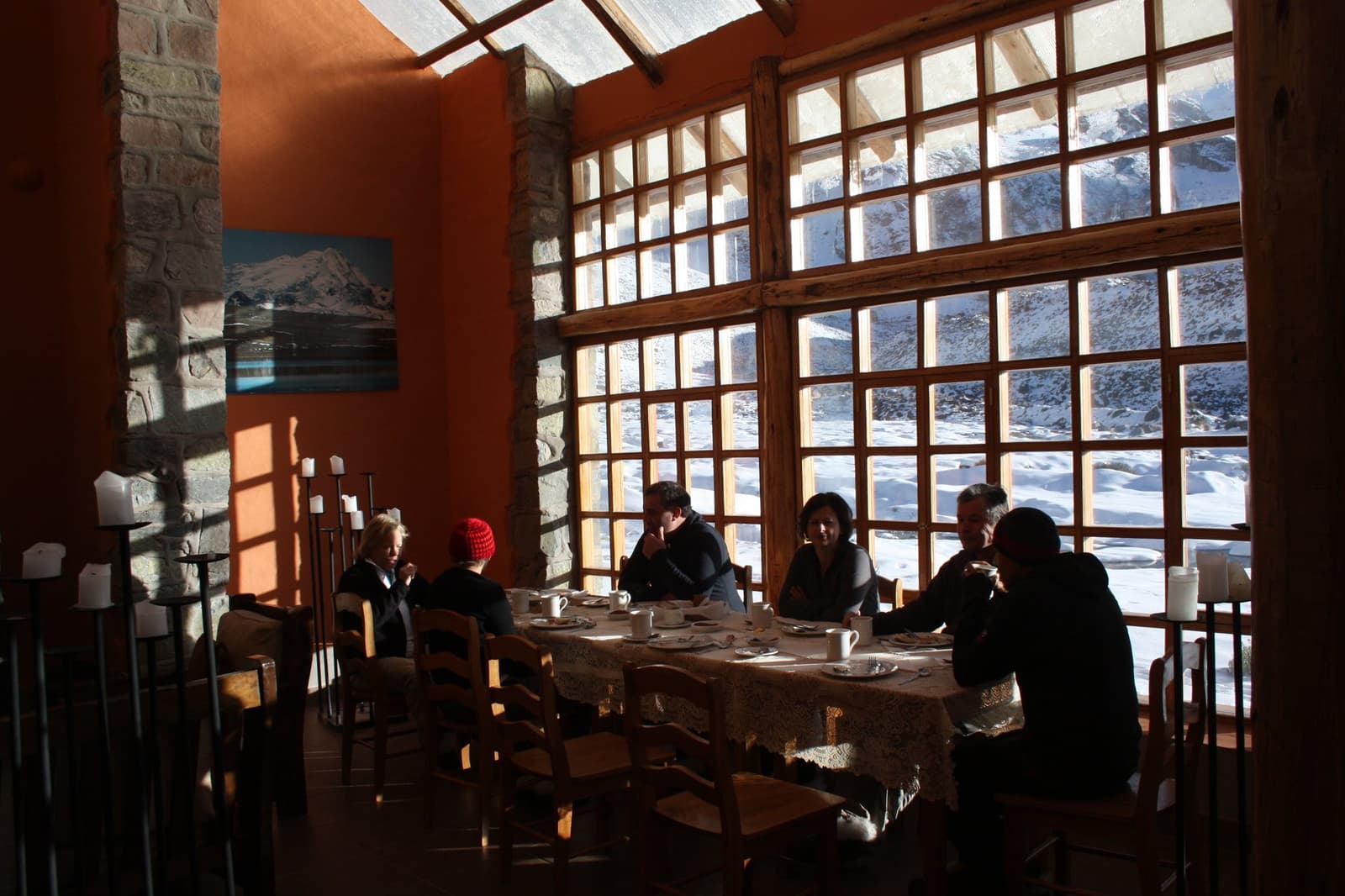Good conversation, hot coffee, snow on the other side of the glass. Travelers sharing breakfast around a long table inside a warm adobe lodge on the Ausangate Lodge Trek, with large wooden-framed windows overlooking a snow-covered Andean landscape outside