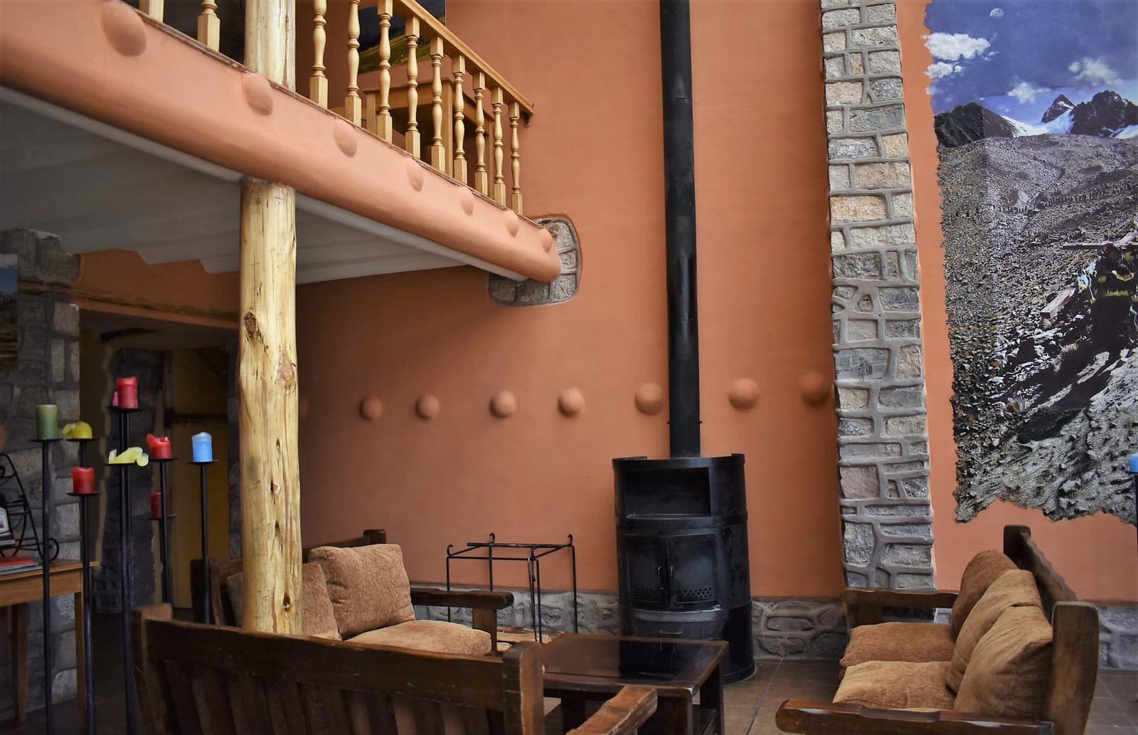 A wood-burning stove after a day above 4,000 m earns its keep. Photo: Rustic lodge common room on the Ausangate Lodge Trek featuring a black wood-burning stove, heavy wooden sofas with tan cushions, a timber-balustrade mezzanine, stone masonry columns, and a large mural photograph of a snowy mountain face on the wall