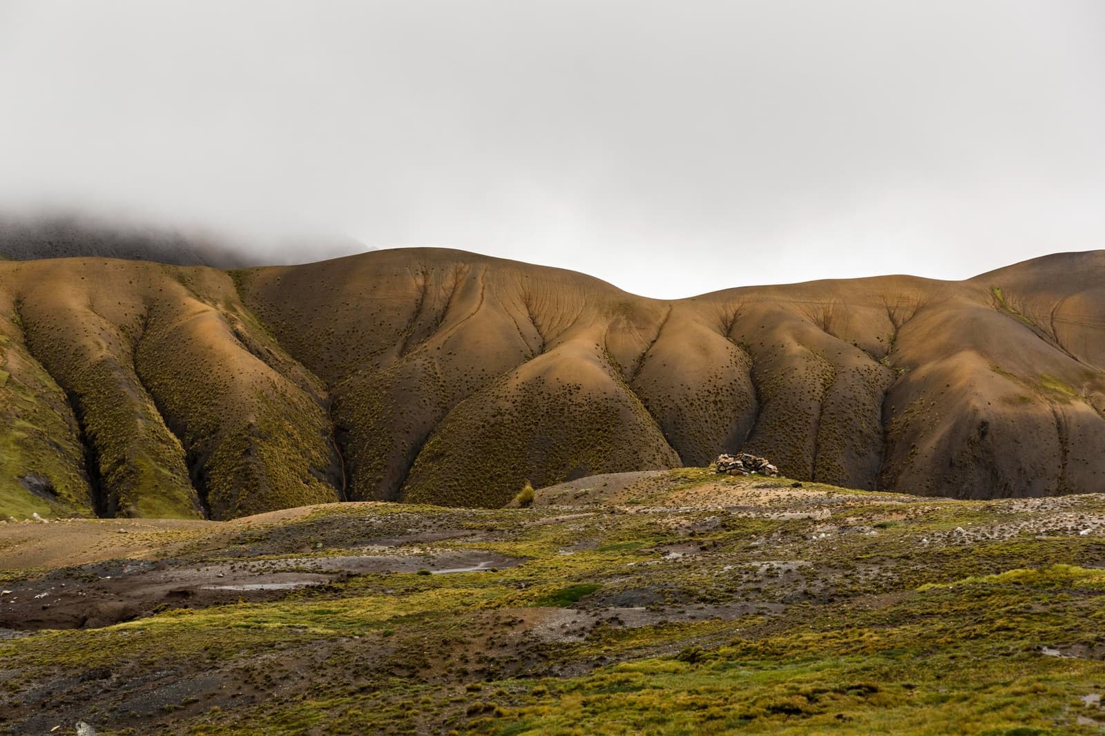 The mountain doesn't do soft edges, but it does do moody. Rounded, erosion-scored hills of the ausangate trek draped in sparse high-altitude vegetation, with low cloud clinging to the ridgeline and a small stone cairn visible on a saddle in the middle ground