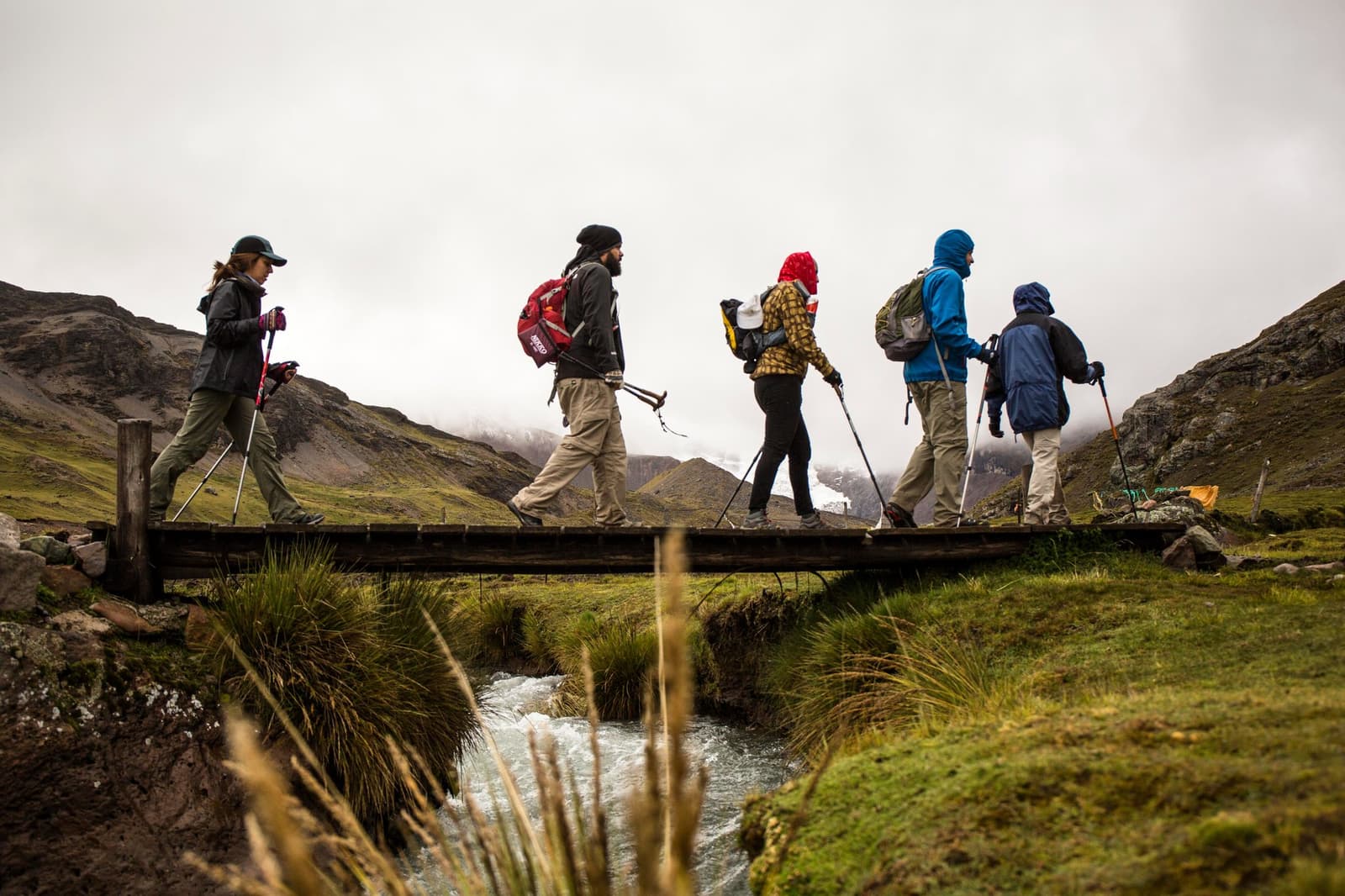 Five people, one plank, and a stream that isn't waiting for anyone. Five hikers with trekking poles and backpacks cross a narrow wooden footbridge over a rushing glacial stream during the Ausangate trek, with cloud-covered Andean peaks and green puna grassland in the background.