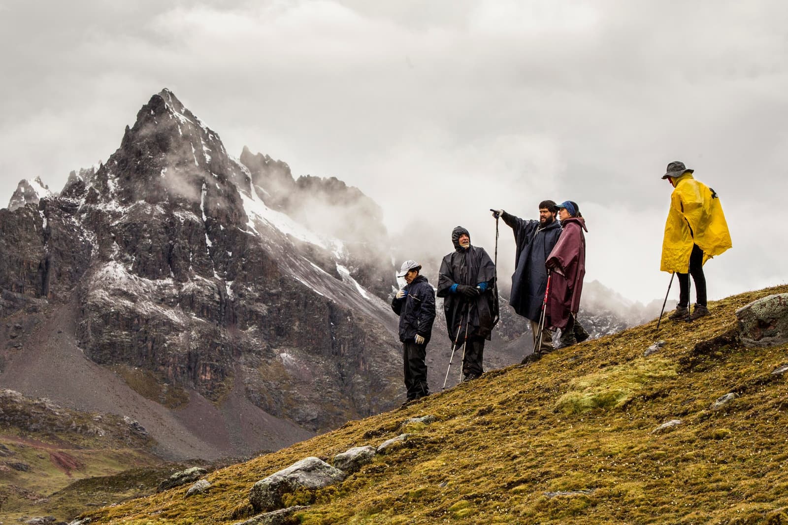Someone always knows which peak is which. Good thing someone brought a finger. Four trekkers in rain gear pause on a mossy high-altitude ridge during the ausangate trek, one pointing toward a jagged snow-dusted peak partially hidden by low cloud