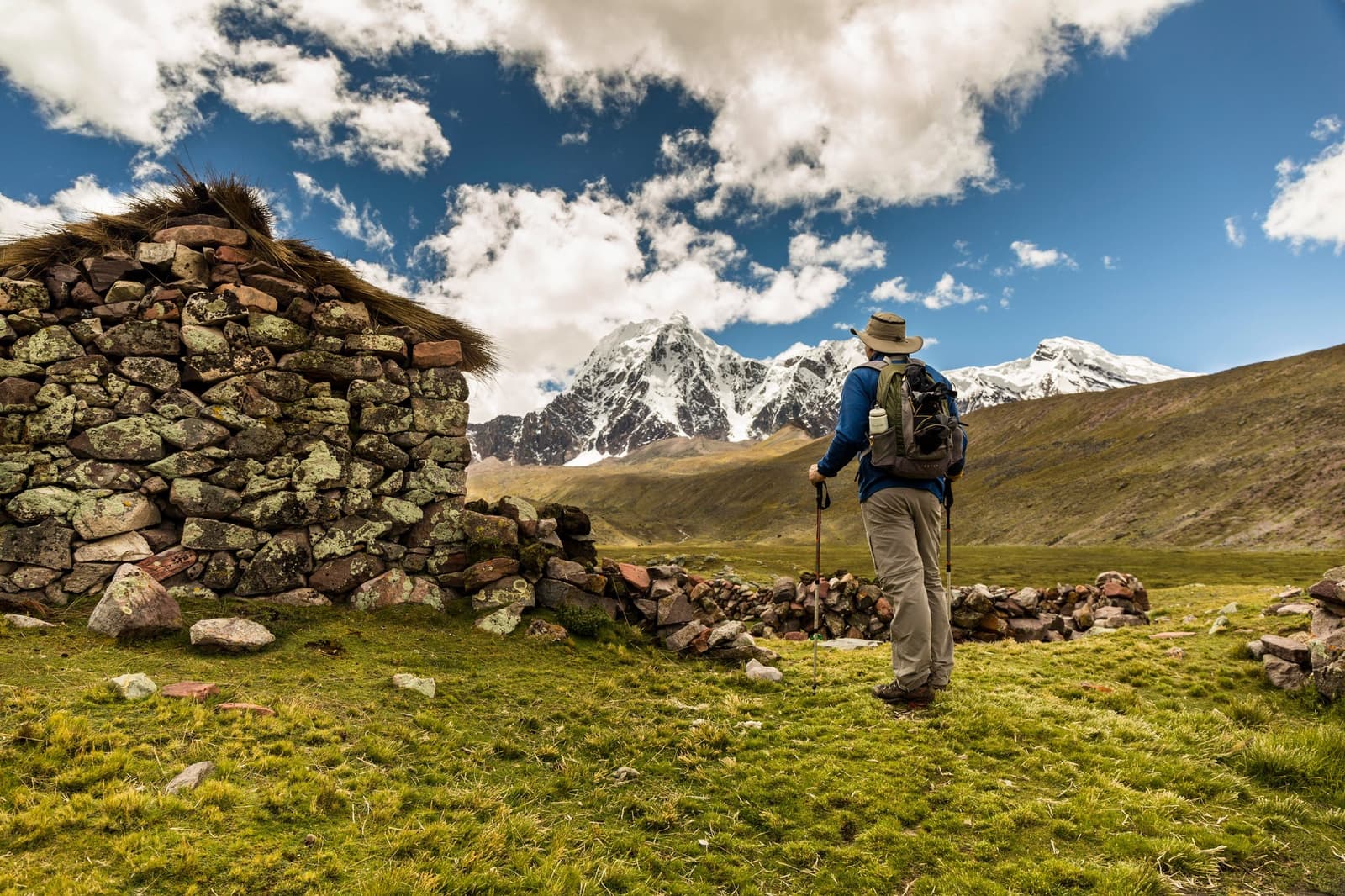 A stone hut older than the trail itself, still earning its keep up here. A hiker with trekking poles and a daypack stands beside a dry-stone shelter with a thatched roof on the ausangate trek, facing a snow-capped mountain peak rising above a green high-altitude valley under a blue sky with clouds