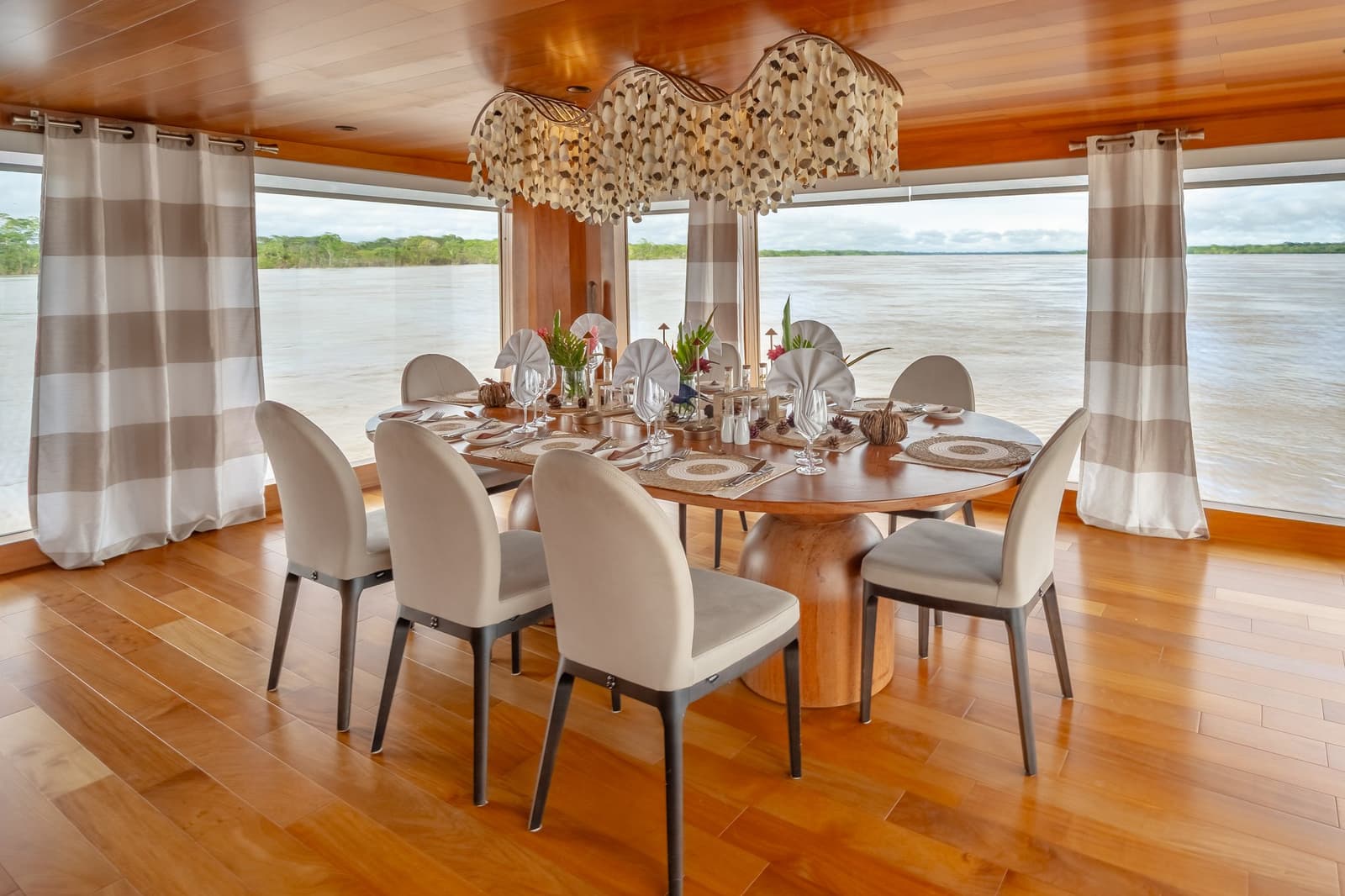 Jantar com o rio ali do lado, sem lista de espera. Photo: Mario Gomi Mesa de jantar oval elegantemente posta com lustre de conchas e vista panorâmica para o rio através de janelas do chão ao teto a bordo do cruzeiro Delfin Amazon