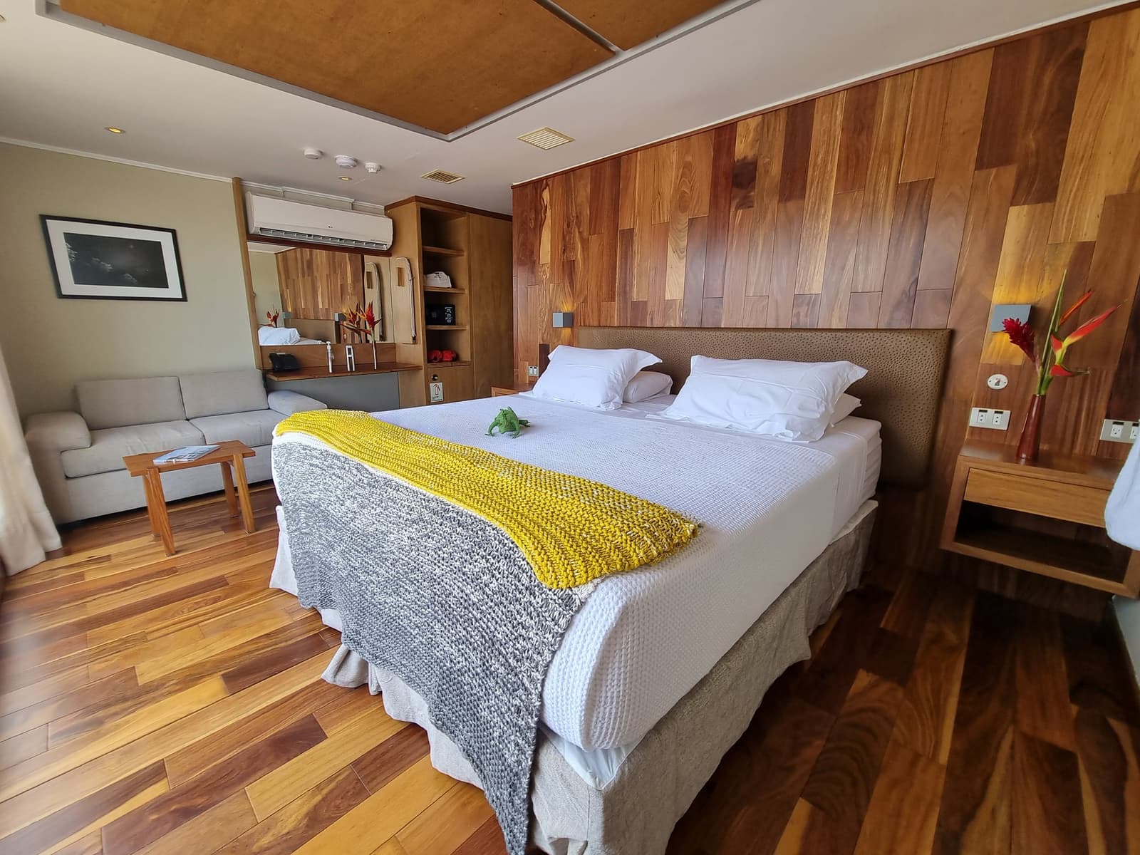 Confortável o suficiente para fazer você repensar o passeio de skiff às seis da manhã. Suite espaçosa do cruzeiro Delfin Amazon com cama de casal em linho branco e manta de tricô amarela, painéis de madeira aquecidos, piso de madeira, área de estar com sofá e uma flor tropical vermelha sobre a mesinha de cabeceira