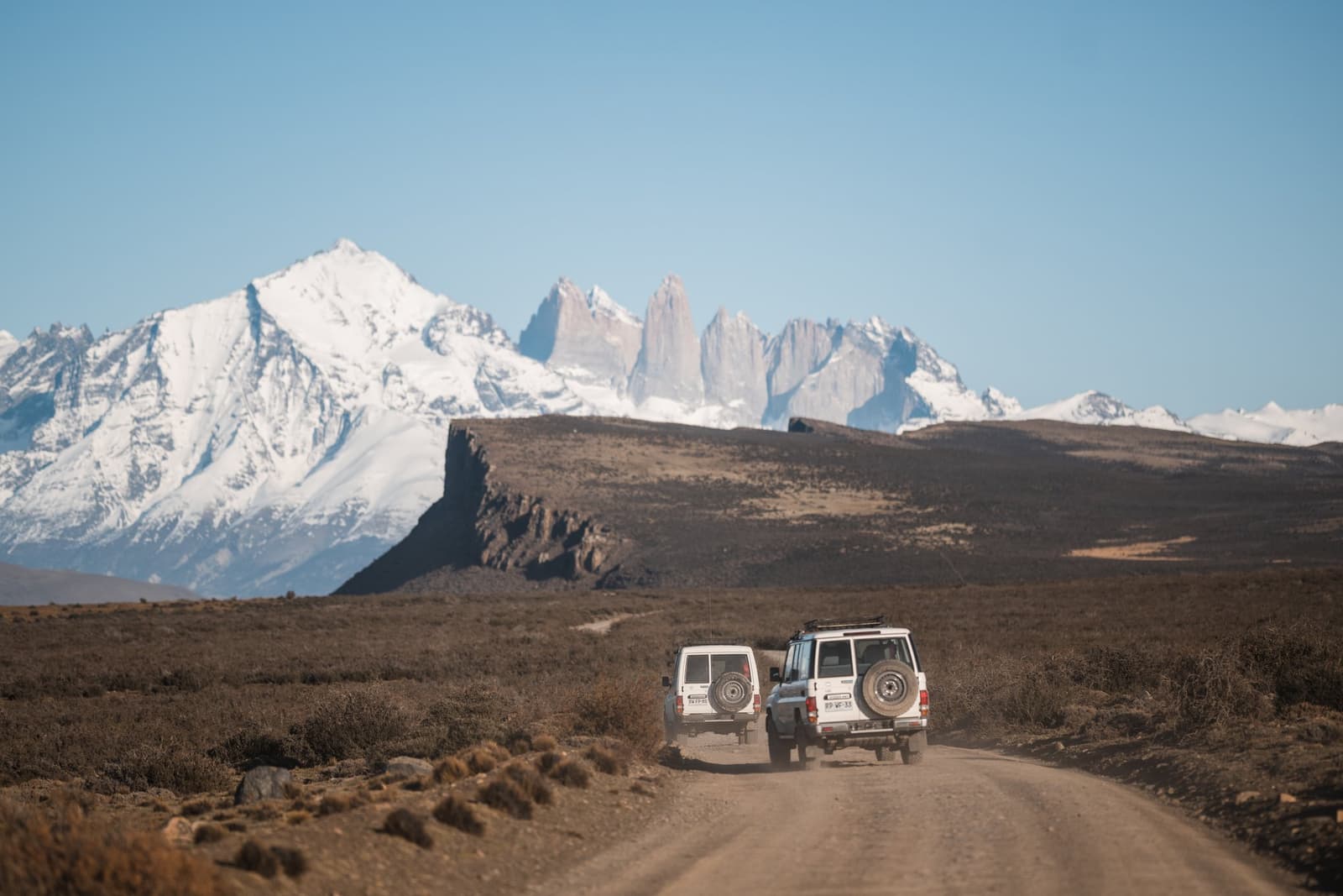 Dust, open steppe, and those towers straight ahead. Some roads earn the drive. Photo: CARLOS DIAZ Two white 4x4 vehicles driving along a dirt road at Estancia Cerro Guido, with the granite towers of Torres del Paine and snow-capped Andean peaks rising across the open Patagonian steppe under a clear blue sky