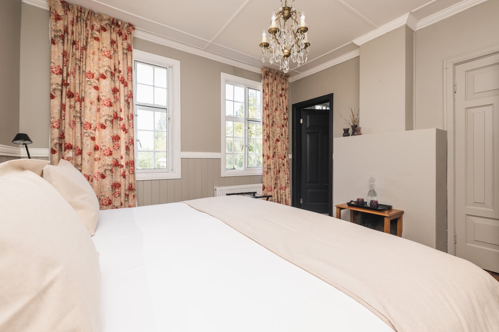 Historic ranch bones, fresh linen, and a chandelier that means business. Photo: CARLOS DIAZ Spacious double room at Estancia Cerro Guido with a crystal chandelier, floral curtains, crisp white bedding, and two sash windows overlooking garden greenery