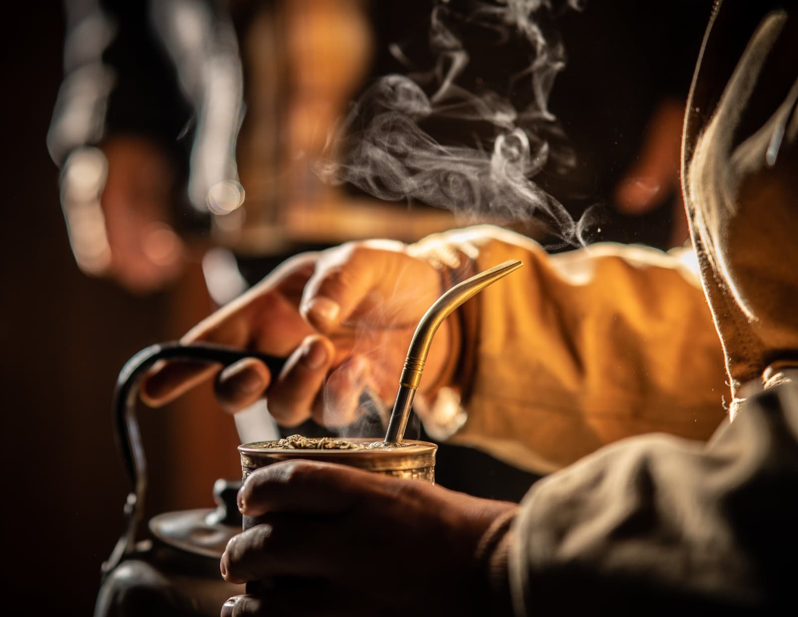 The gauchos' afternoon mate: not a ceremony, just the way things are done. Weathered hands cupping a small mate gourd with a gold bombilla, steam rising in warm light at estancia cerro guido, with a kettle visible in the background