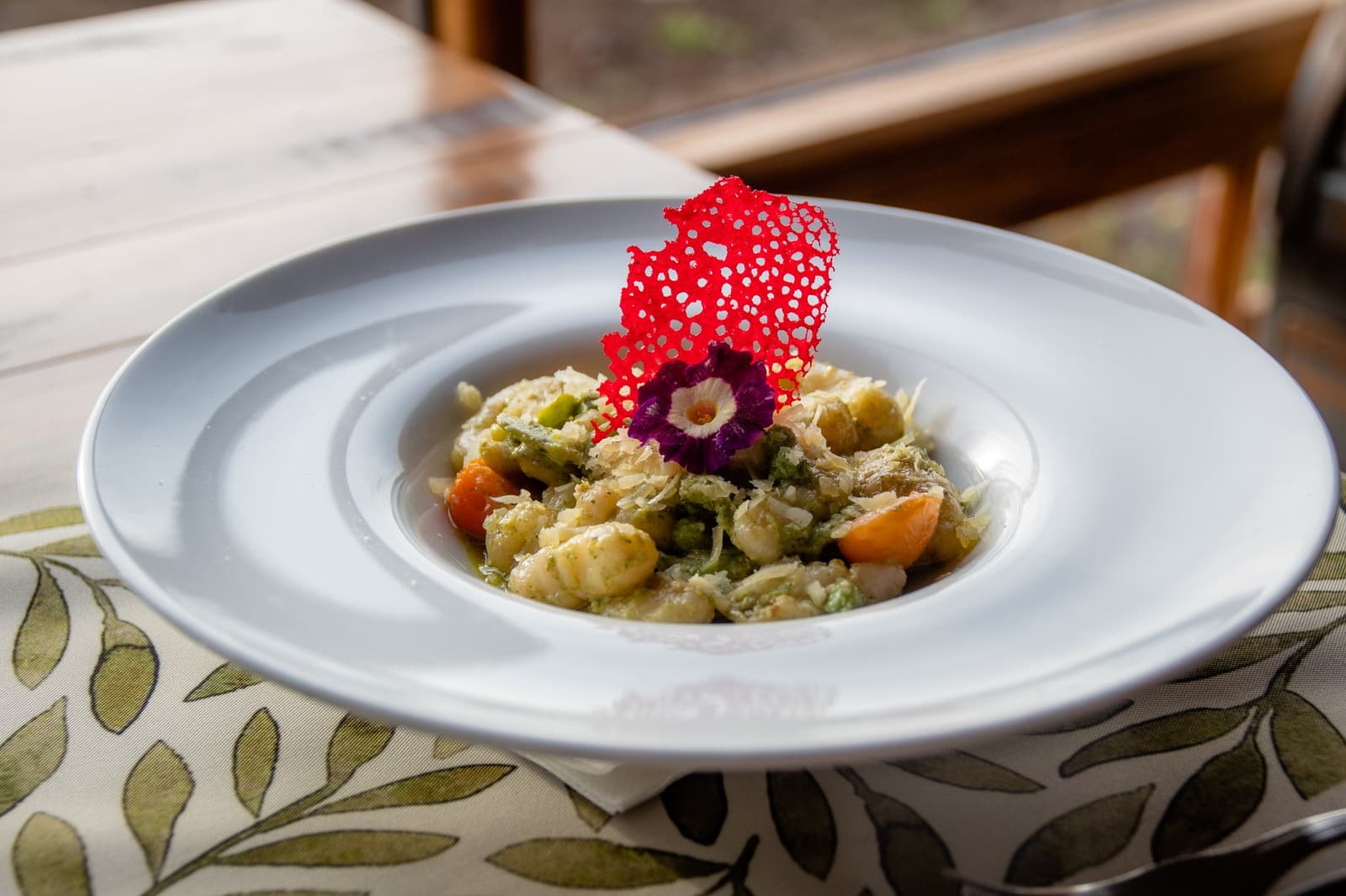 Patagonian wind outside, a tuile and an edible flower inside. The kitchen has opinions. A plated gnocchi dish at Estancia Cerro Guido, garnished with peas, cherry tomatoes, shaved cheese, an edible purple flower, and a lacy red tuile, served on a white wide-rim plate atop a botanical-print table runner