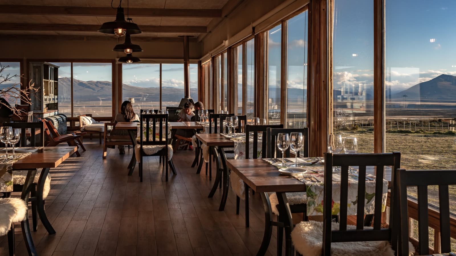 Lunch tastes different when the steppe is your tablecloth. Wood-paneled dining room at Estancia Cerro Guido with set tables, sheepskin chair cushions, and floor-to-ceiling windows overlooking the open Patagonian steppe and distant mountains under a clear blue sky