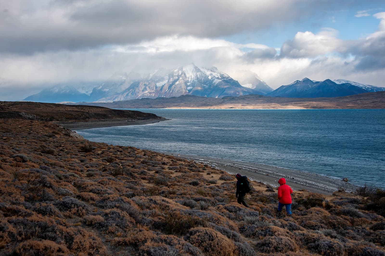 Cold wind, big mountains, good company. Some days earn their dinner. Two hikers in winter jackets walk along a gravel lakeshore at Estancia Cerro Guido, with snow-capped Patagonian peaks emerging from dramatic cloud cover across the blue lake
