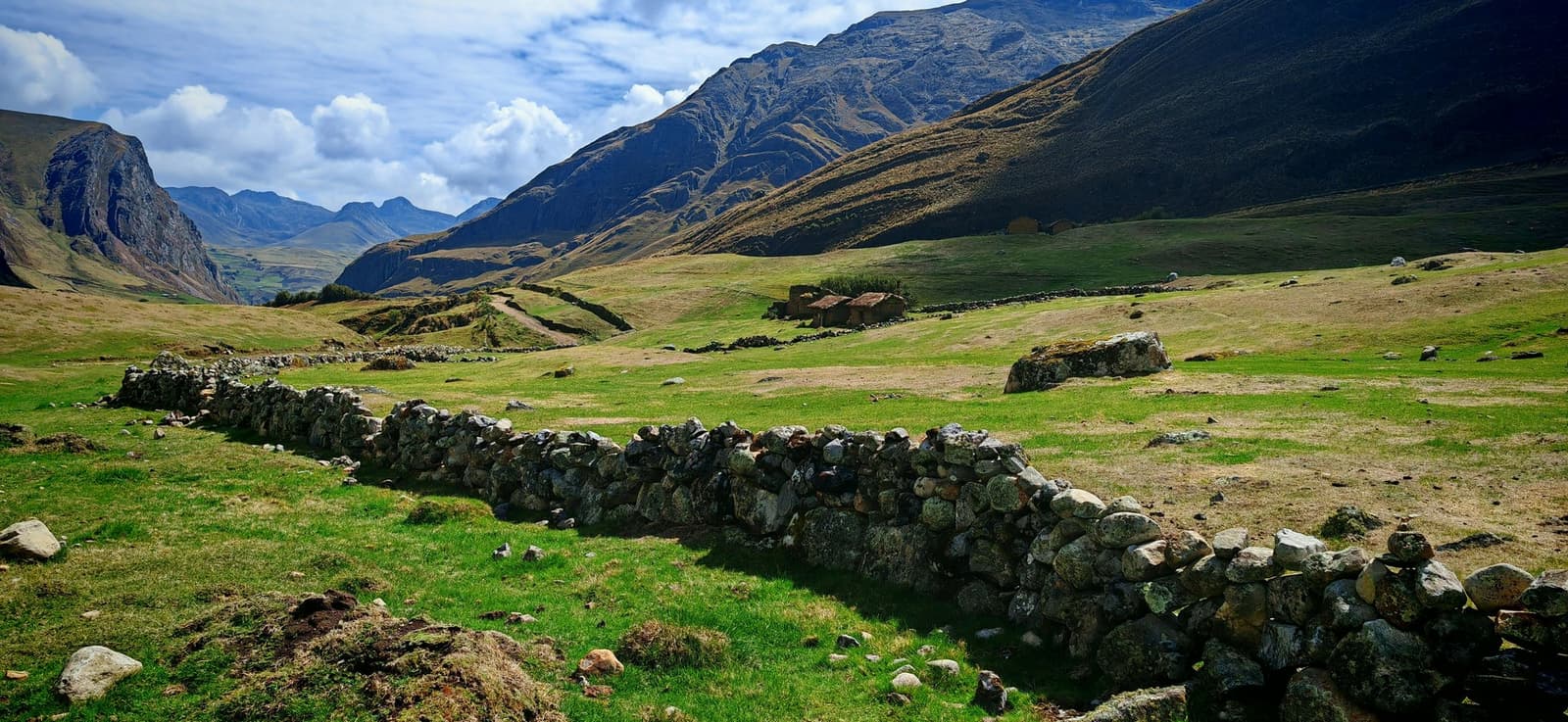 Somebody built these walls long before trekking was a thing people did for fun. Ancient dry-stone walls cross a broad green high-altitude valley on the Huayhuash circuit, with a ruined stone structure in the middle distance and steep Andean peaks rising under a partly cloudy sky