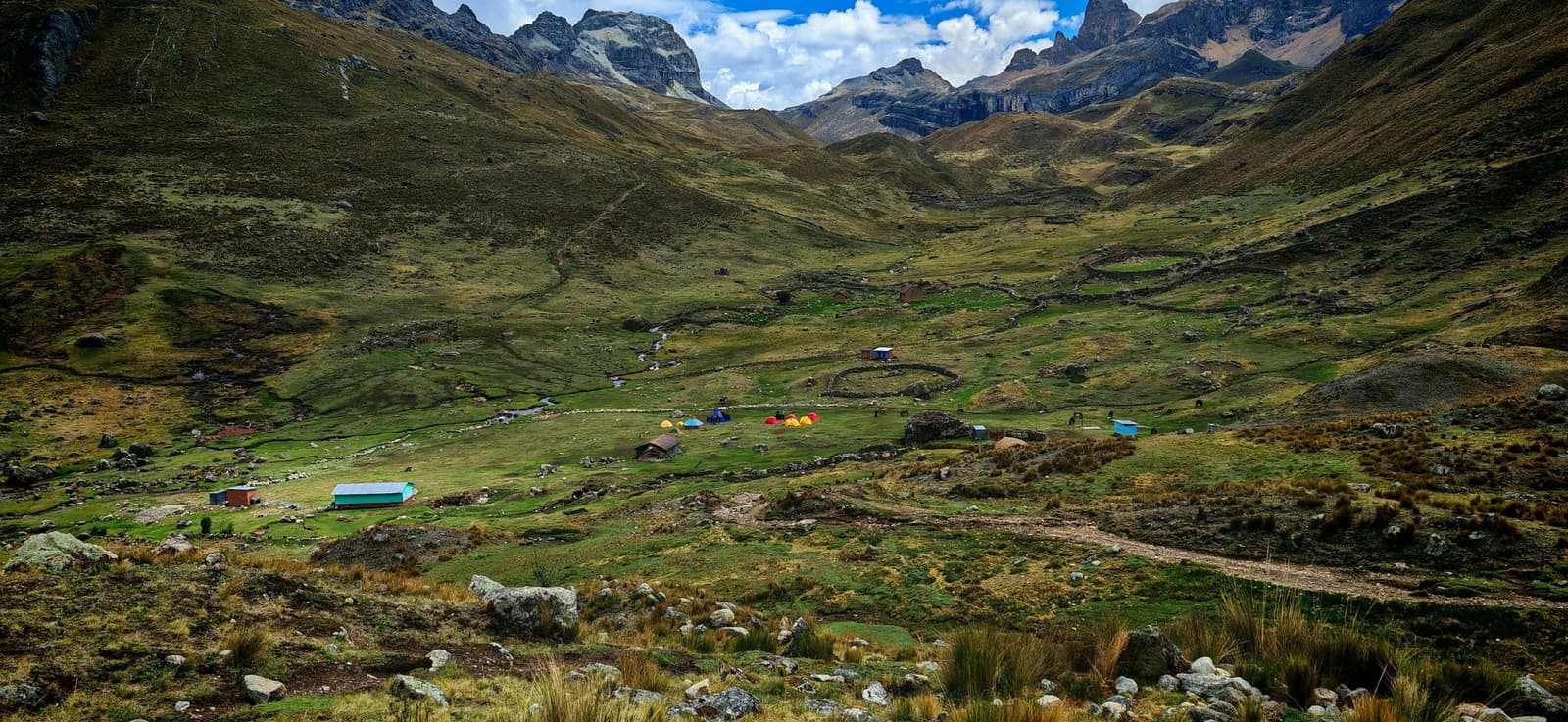 Tiny tents, big mountains, and a stream that doesn't know how far from anywhere it is. A wide Andean valley on the huayhuash circuit showing a small trekking camp with colorful tents and stone-walled enclosures on green puna grassland, surrounded by rugged rocky peaks under a partly cloudy sky