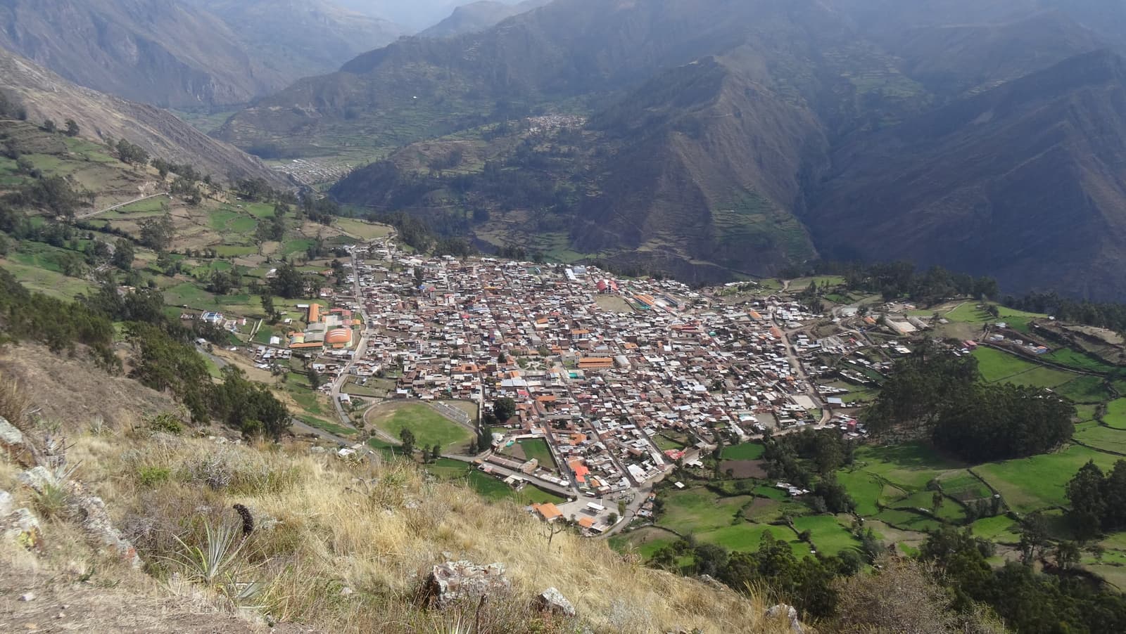 A whole town tucked into the folds of the Andes, proof the mountains have always made room for people. Aerial view of a compact Andean town nestled in a steep mountain valley on the huayhuash trek route, with green terraced fields and dry rocky slopes surrounding the settlement