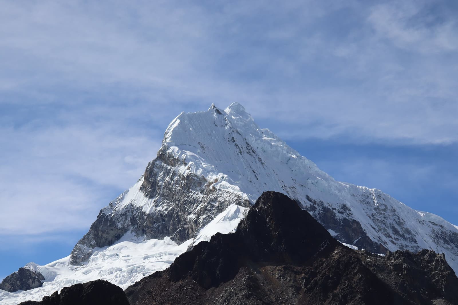 Tem cume que não precisa de legenda para te deixar sem fôlego. Um pico de alta altitude densamente glaciado na trilha Huayhuash, com cume pontiagudo e faces cobertas de gelo se erguendo contra um céu azul com fina cobertura de nuvens acima de cristas rochosas escuras
