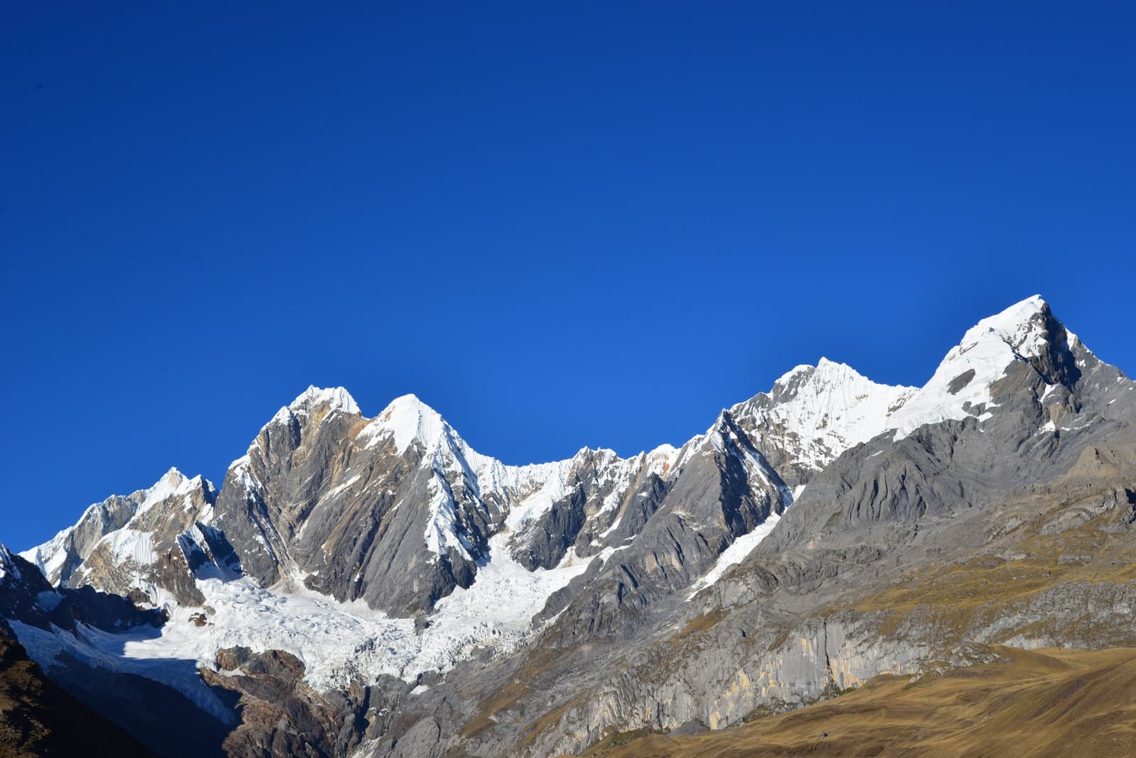 Clean air, sharp ridgelines, and not a cloud willing to compete. Snow-capped rocky peaks of the Huayhuash trek range rising sharply against a deep blue sky, with glaciers clinging to the steep grey faces and dry golden slopes in the foreground
