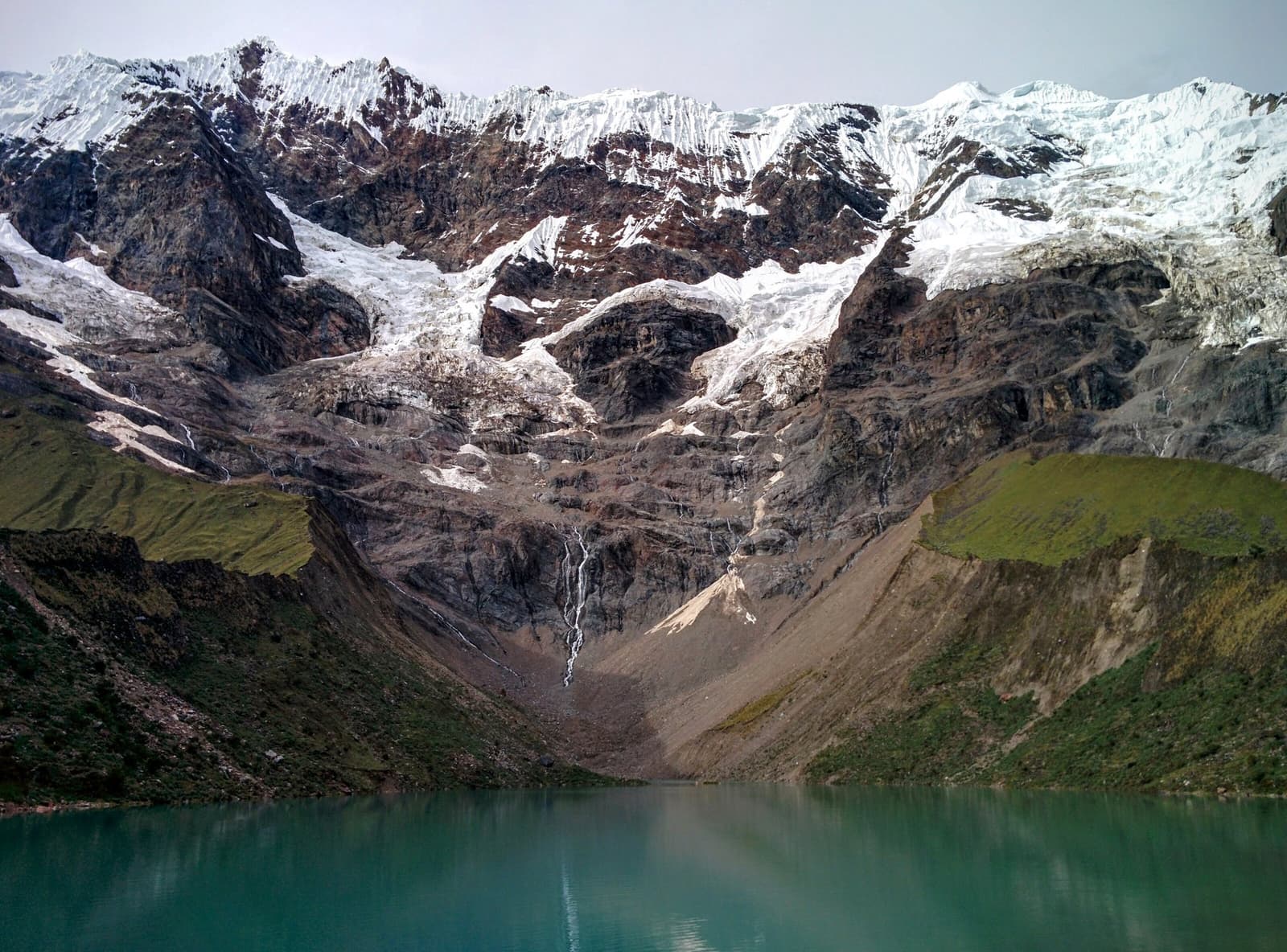 A neve vira fio d'água, o fio vira lago, e o lago fica ali com aquela cor impossível. Águas turquesa glaciais do lago Humantay no trek Salkantay de 5 dias, com uma parede rochosa coberta de neve e fios de cachoeira descendo em direção à margem