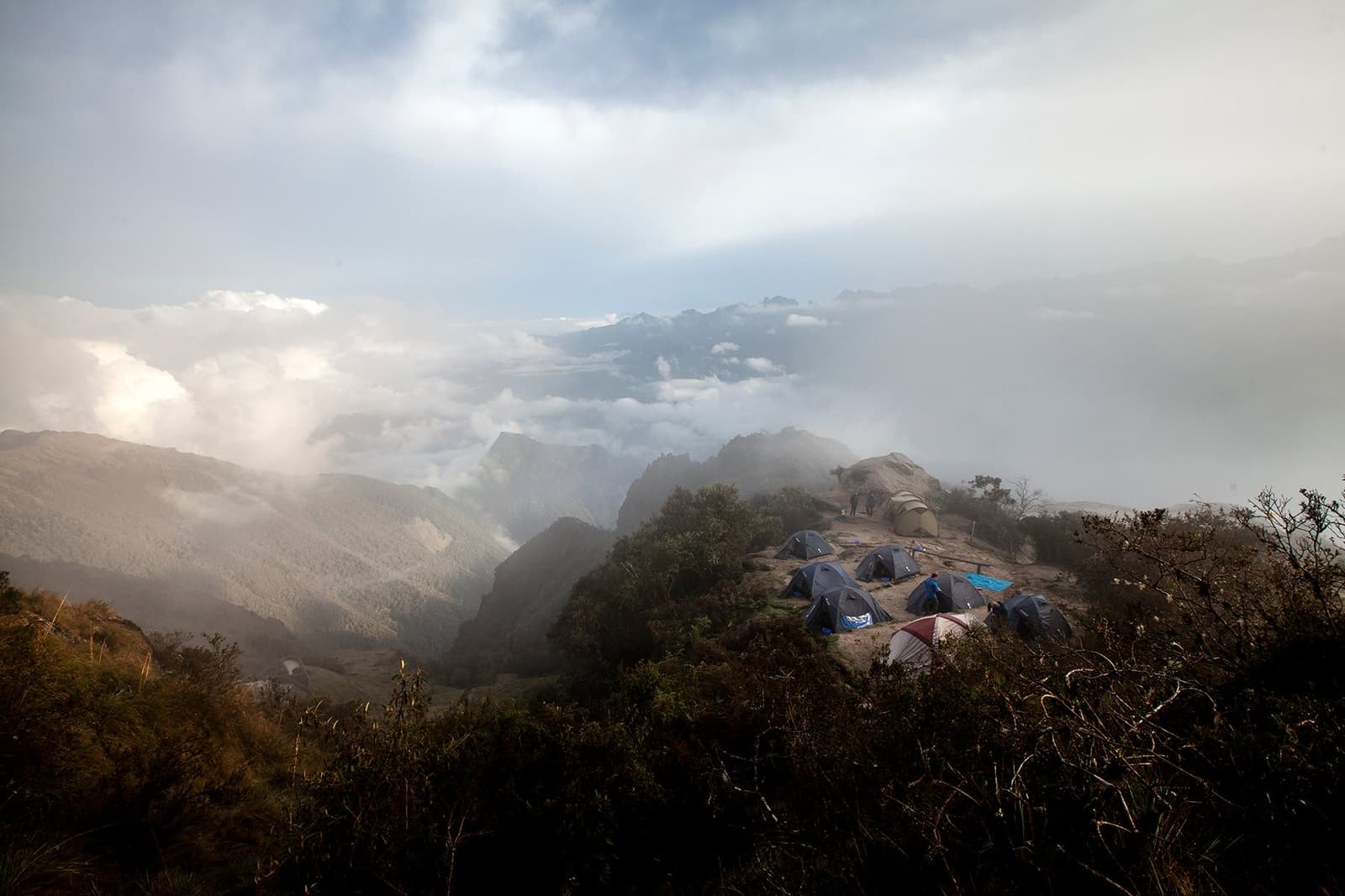 The clouds came up fast. The tents held their ground. A cluster of dome tents pitched on a rocky ridge along the Inca Trail to Machu Picchu, surrounded by mountain scrub and a thick sea of clouds filling the valleys below, with distant peaks emerging above the cloud layer