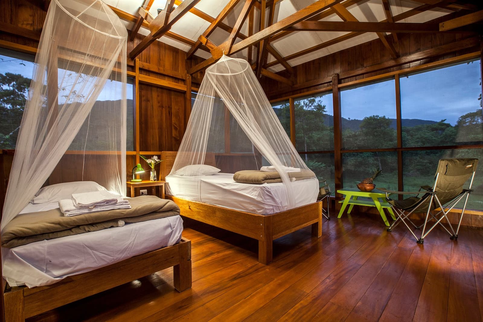 Nets up, forest on three sides, river just below. Sleep comes easy here. Twin beds with white mosquito-net canopies on a hardwood floor inside a wooden cabin lodge, with panoramic windows looking out to forested hills at dusk — overnight accommodation on a Machu Picchu Amazon tour that crosses Peru by land.