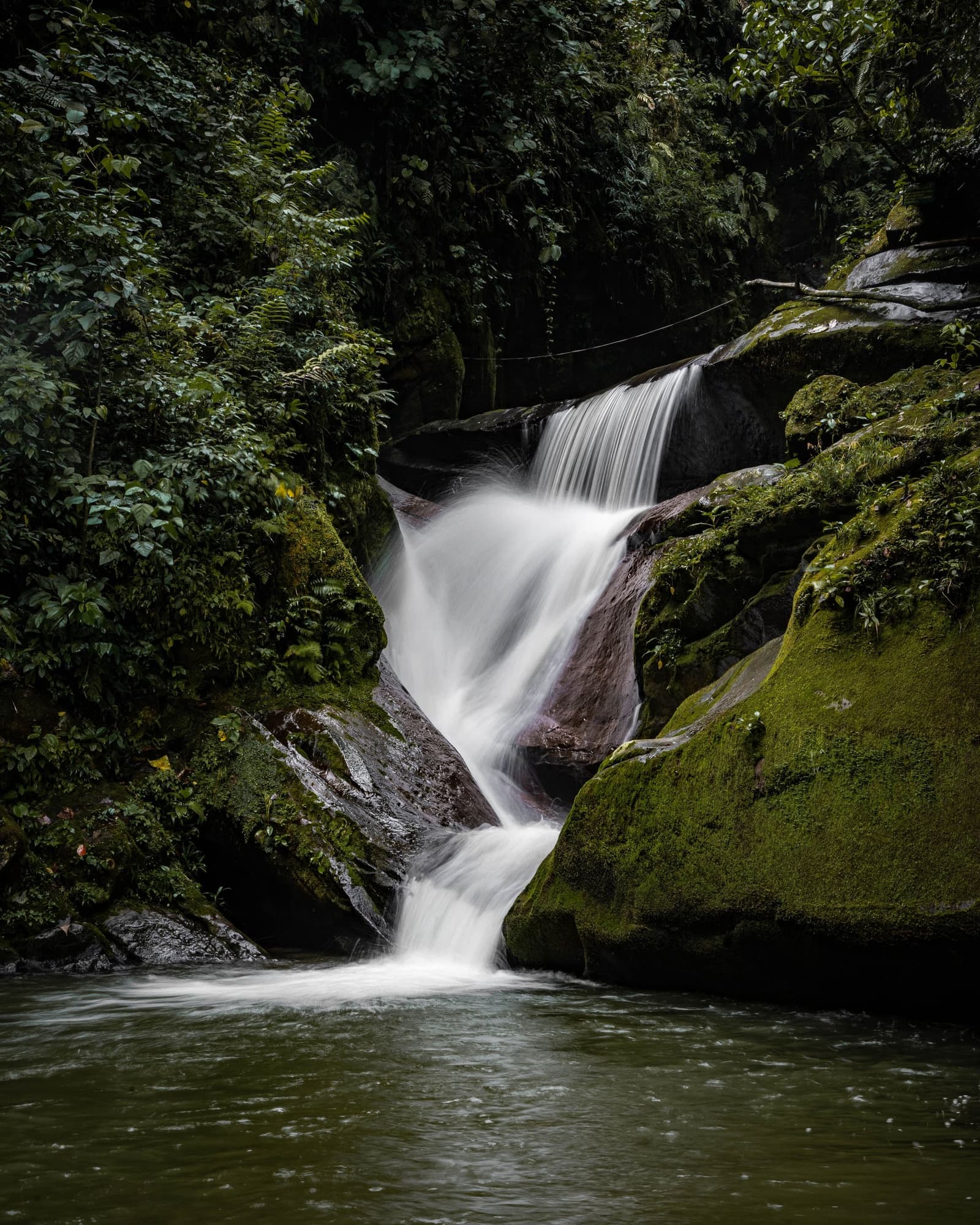 Cold, clear, and completely indifferent to your schedule. A silky long-exposure waterfall cascades over dark moss-covered boulders into a calm pool, surrounded by dense cloud forest vegetation on the machu picchu amazon tour overland route through Peru