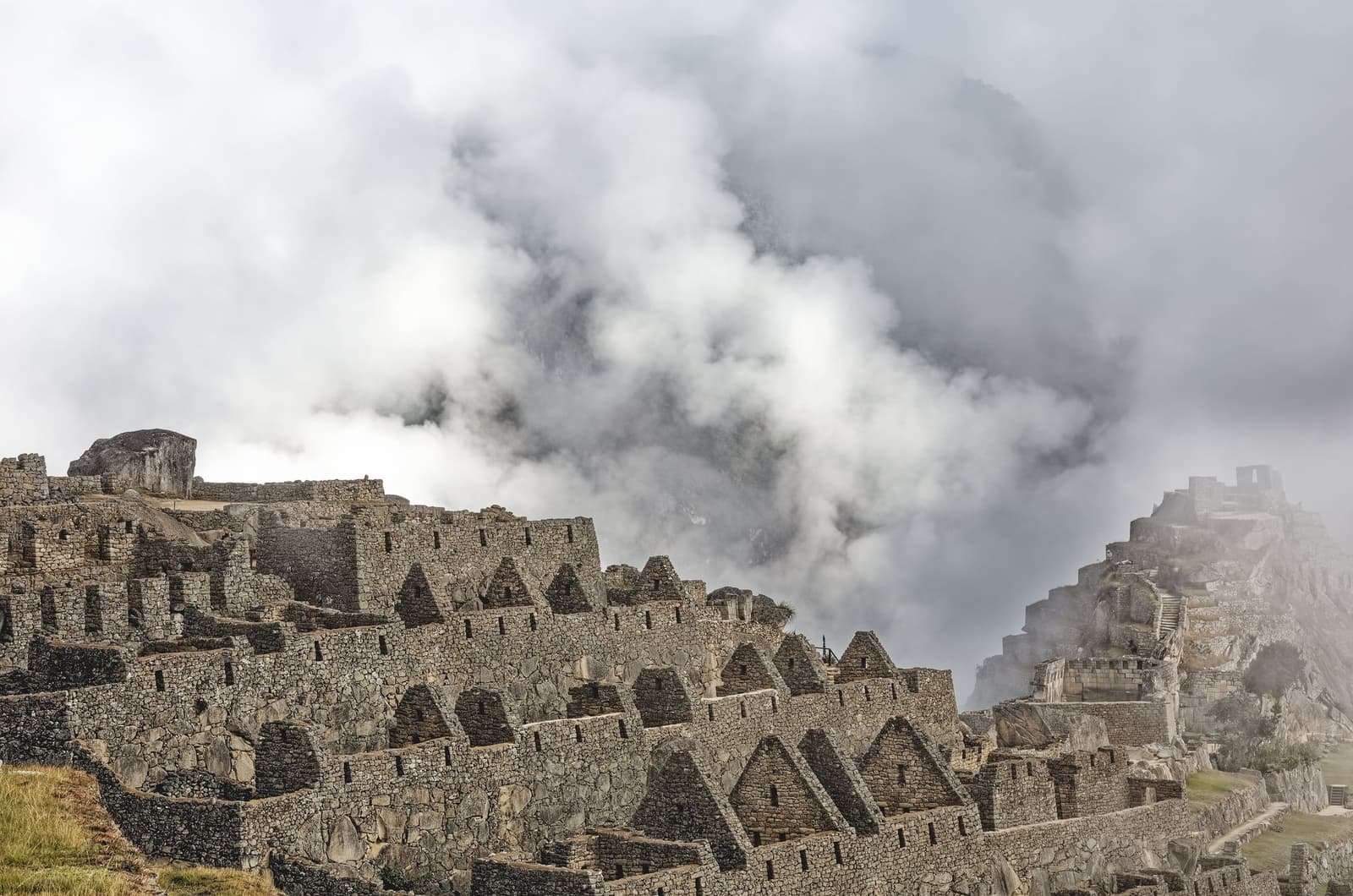 Cloud rolls through on its own schedule, and the stones don't mind at all. Rows of roofless Inca stone structures at Machu Picchu shrouded in thick cloud on a machu picchu amazon tour, with terraced walls and trapezoidal doorways descending the ridge and a distant hilltop structure emerging from the mist