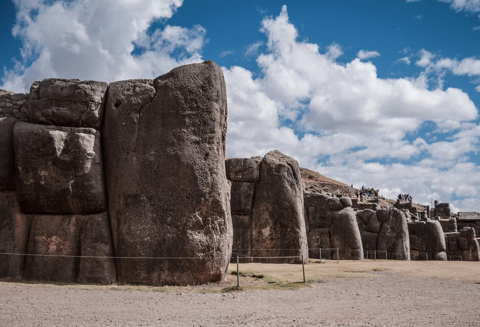 Stones so large the Inca never needed mortar, and still haven't. Massive precisely fitted limestone blocks of the Sacsayhuaman fortress walls rise against a blue sky with white clouds, with small groups of visitors visible on the terraces in the distance, a stop on this machu picchu amazon tour