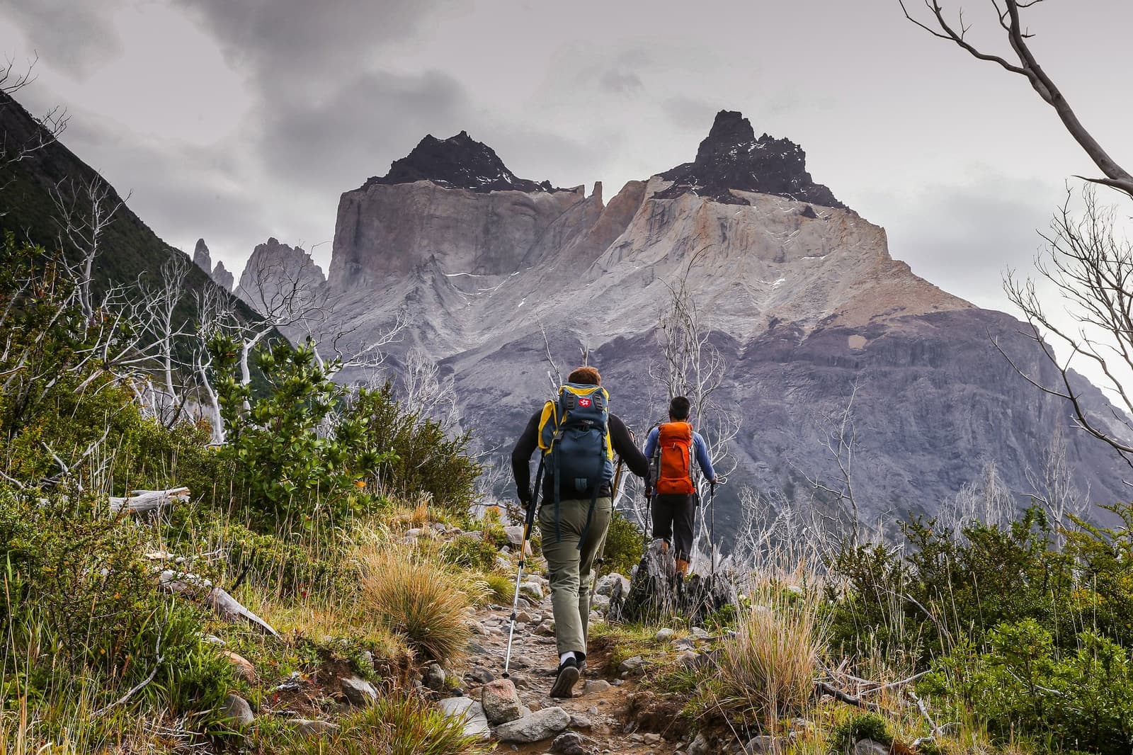 The mountains don't wait for perfect weather, and neither should you. Photo: WWW.SOTOFOTOS.COM Two hikers with large backpacks and trekking poles walk a rocky trail on the self-guided O Circuit, with the granite walls and dark horns of the Cuernos del Paine rising steeply under a cloudy sky, surrounded by scrubby vegetation and bleached dead trees