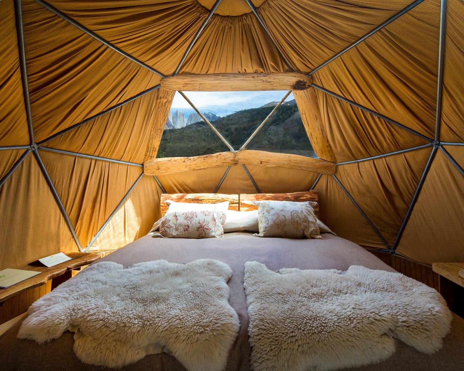 Sheepskin, a wood-framed bed, and the Towers right there in the window. Photo: Standard Dome Interior of a geodesic dome at EcoCamp Patagonia on the O trek in Patagonia, showing a double bed layered with sheepskin throws, patterned pillows, a rustic wood headboard, and a triangular window framing the granite spires of Torres del Paine in the distance