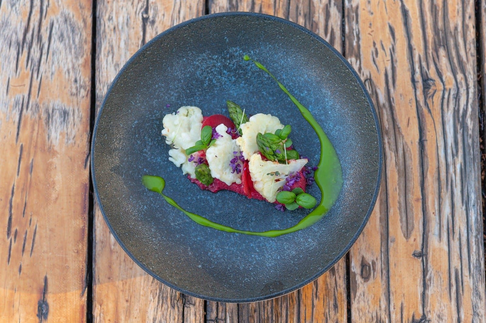 After big days on the circuit, this is what a proper sit-down meal looks like. Overhead view of a plated vegetable dish on a dark ceramic bowl at EcoCamp Patagonia, featuring roasted cauliflower, asparagus, red beet purée, green herb sauce, and edible purple flowers on a rustic wood table