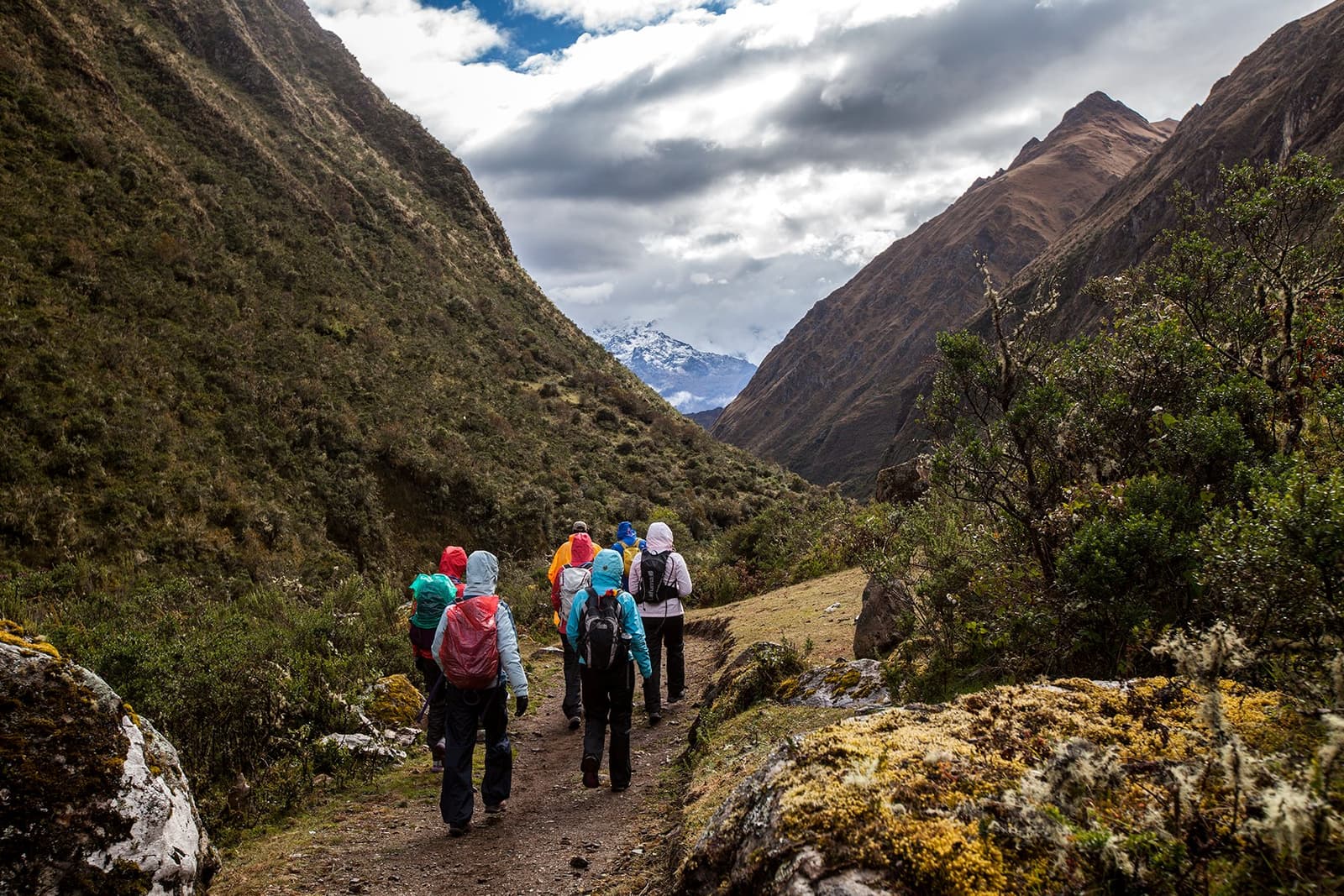 A snow cap at the end of the valley, and nobody else on the trail. That's the point. A group of trekkers in colorful rain jackets walks along a narrow path through a high Andean valley on the off-the-beaten-path Salkantay trek, with a snow-capped peak visible through the clouds at the end of the gorge