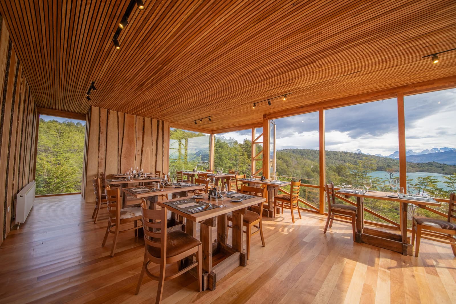 Lunch with that view, and suddenly the hike feels worth every step. Photo: Patagonia Camp Patagonia Camp's wood-lined restaurant with set tables and floor-to-ceiling windows overlooking a turquoise lake and snow-capped peaks beyond the native forest