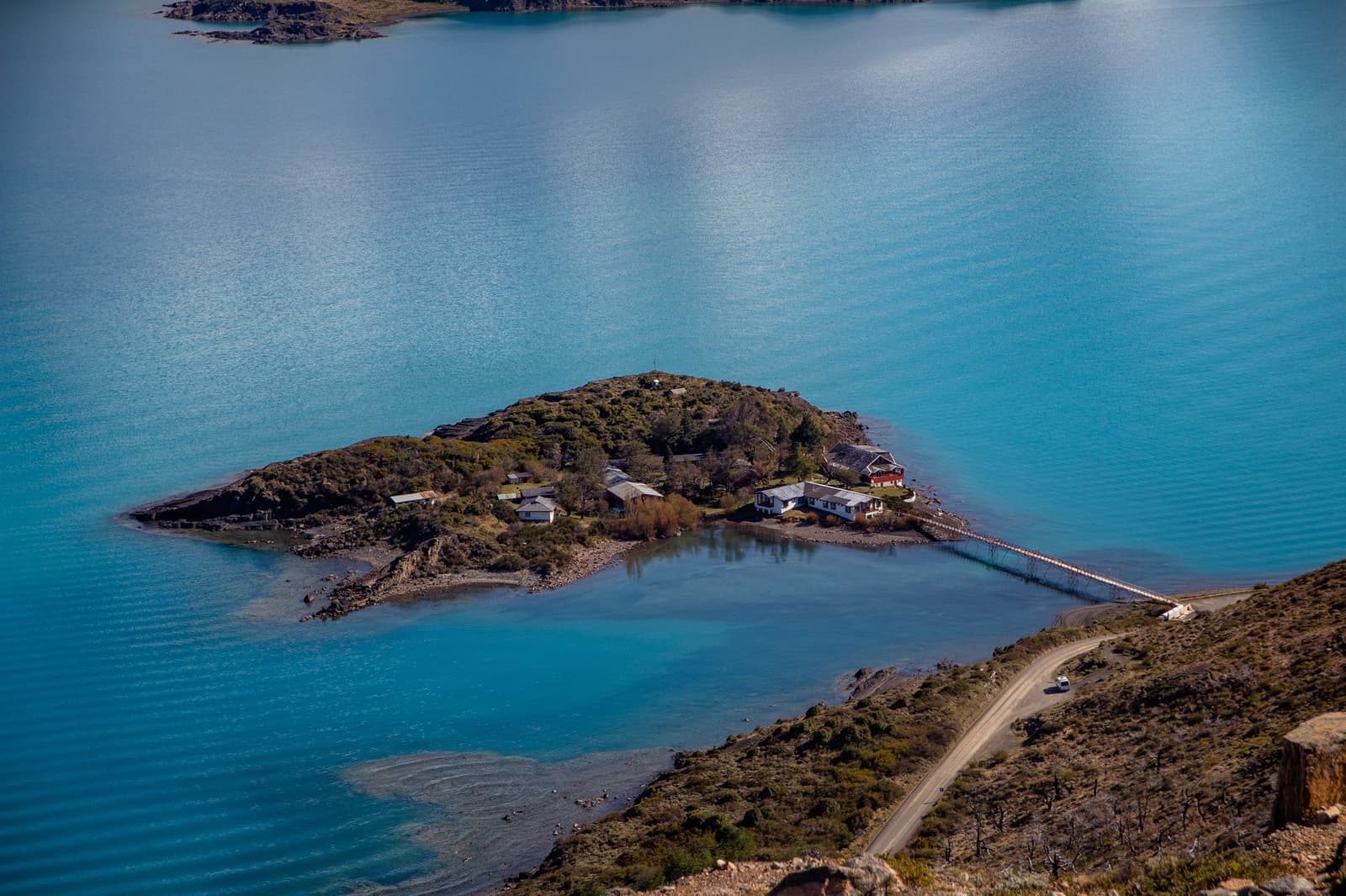 A whole island for a handful of buildings. Lake Toro has a way of making ambition look modest. Photo: Patagonia Camp Aerial view of a small island on turquoise Lake Toro, with several buildings connected to shore by a narrow causeway and a winding gravel road beside the patagonia camp property