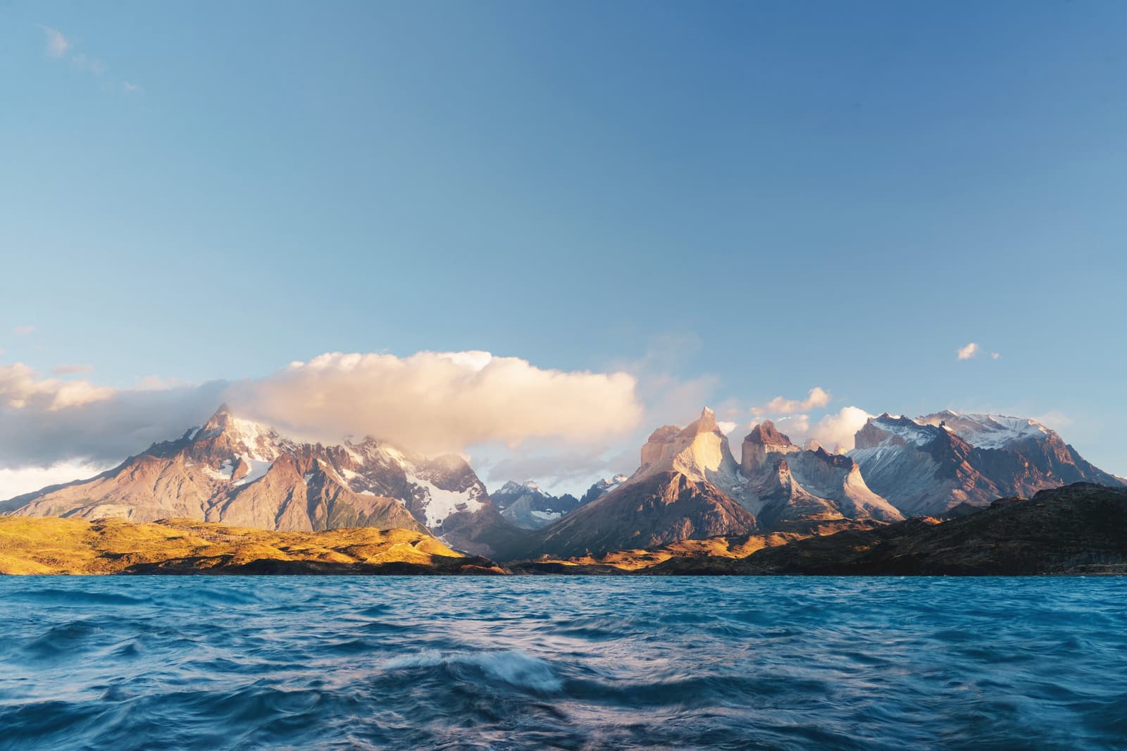 Big mountains, bigger water, and somehow the light always shows up on time. Photo: Patagonia Camp The jagged granite peaks of the Paine Massif rise above golden foothills and deep blue choppy water, seen from lake level at Patagonia Camp on Lake Toro under a clear sky with soft clouds