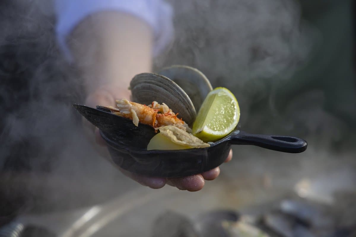 Steam still rising, lime at the ready. Some meals just don't wait. Photo: Patagonia Camp A hand holds a small cast-iron skillet of steaming clams, a shrimp, and lime wedges at Patagonia Camp, smoke rising around the dish outdoors