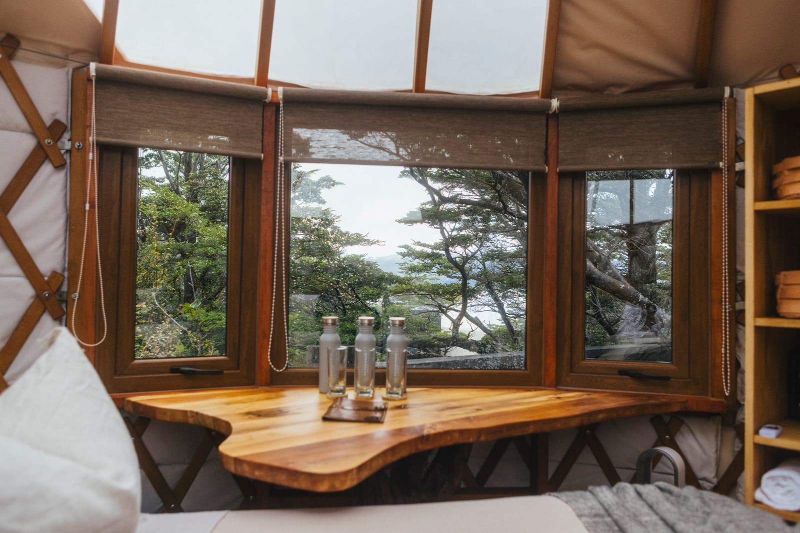 The forest outside, a warm wood table in front. Some rooms earn their keep. Photo: Patagonia Camp Inside a Patagonia Camp yurt, a live-edge wooden table with reusable water bottles sits beside panoramic curved windows looking out over native lenga forest