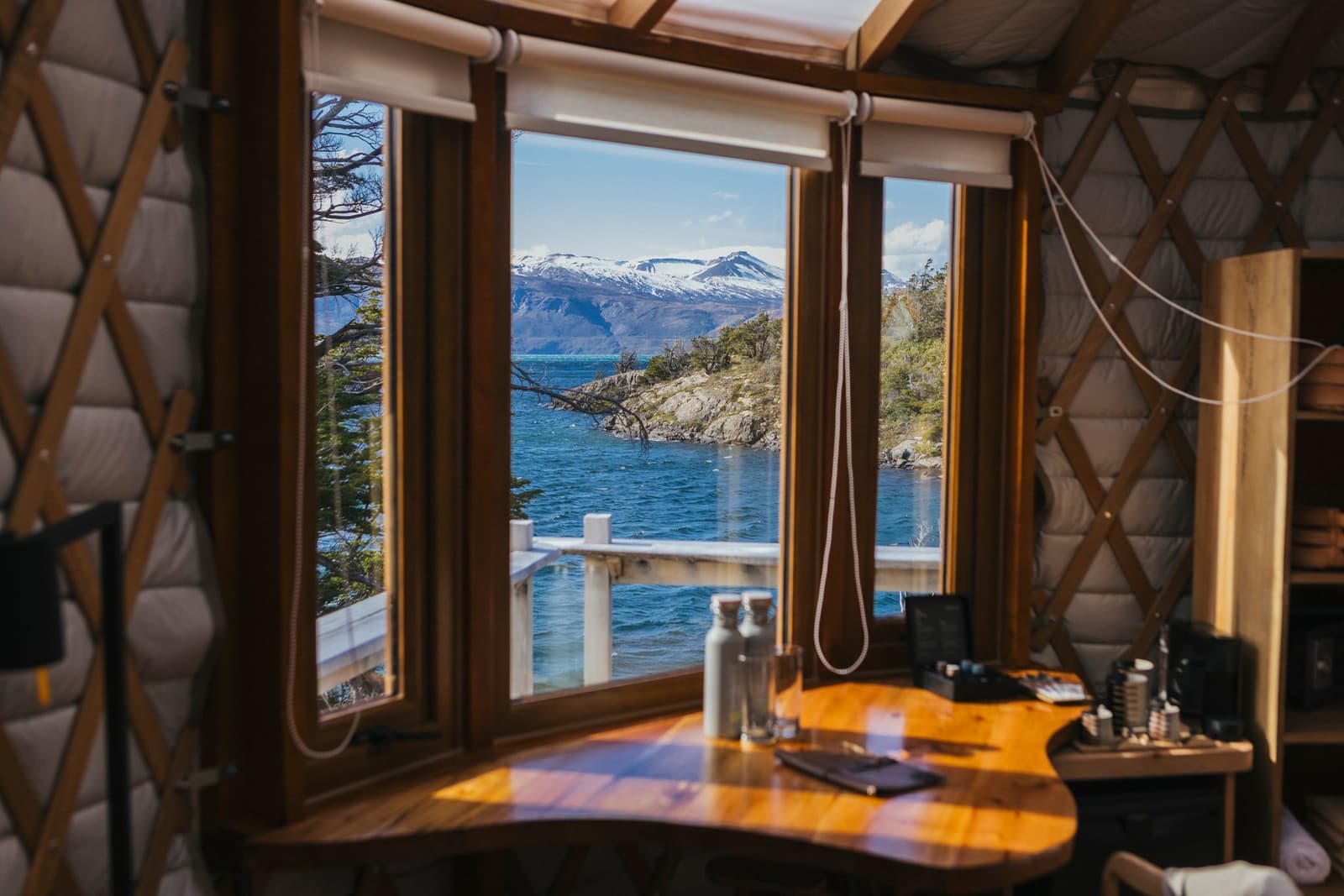 That desk isn't for working. It's for staring at the lake until lunch. Photo: Patagonia Camp Interior of a patagonia camp yurt with a curved wooden desk and water bottles in the foreground, large framed windows opening onto a deck railing, deep blue lake waters, a rocky tree-covered peninsula, and snow-capped mountains under a sunny blue sky