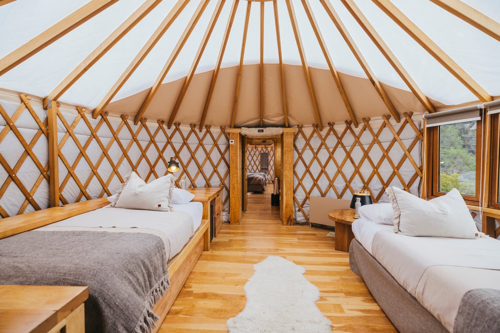 Plenty of room, and the round walls mean everyone gets a good corner. Photo: Patagonia Camp Interior of a patagonia camp yurt with two beds on wooden frames, white linen, a sheepskin rug on hardwood floors, and a latticed wood-and-canvas circular wall structure leading to a second connected yurt