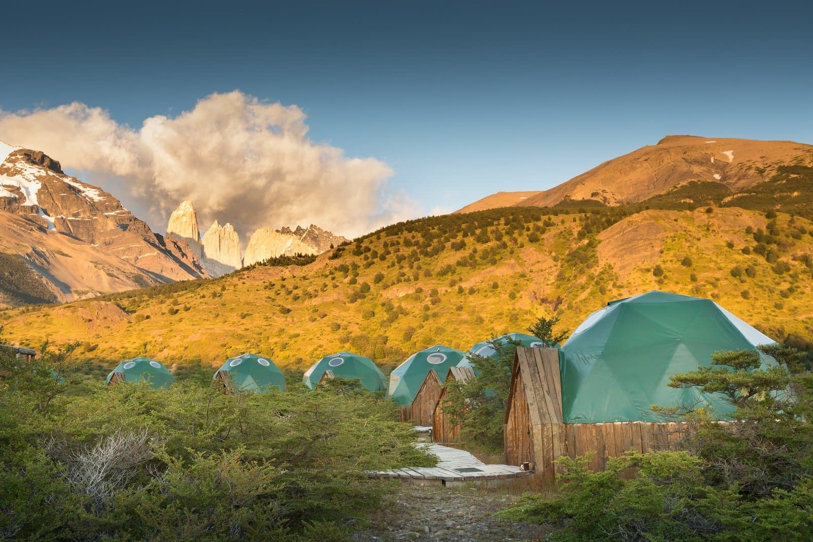 Your home between puma searches: small domes, big views, wood stove waiting. A row of teal geodesic dome accommodations at EcoCamp Patagonia, set among native scrub with the iconic granite towers of Torres del Paine lit by warm golden light in the background