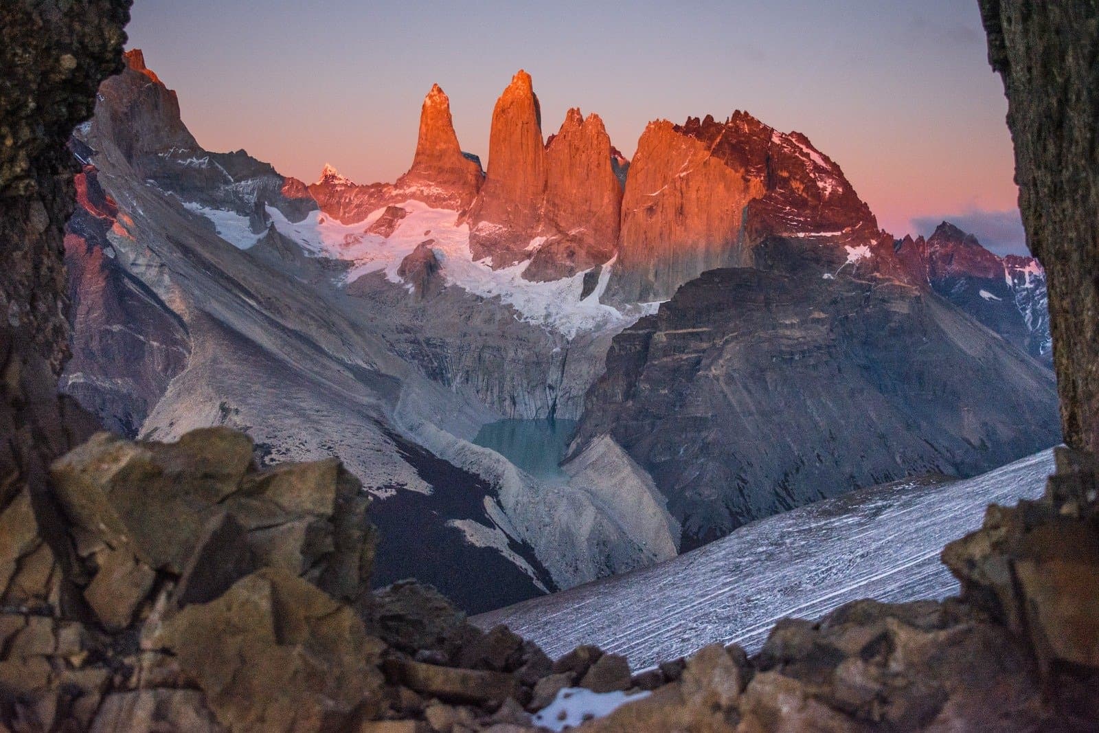 First light on the towers, and the park has already been awake for hours. The three granite towers of Torres del Paine glowing deep orange at sunrise, framed by rock formations in the foreground, with a glacial teal lake nestled below the peaks and snow-streaked slopes — the dramatic landscape at the heart of puma trekking in Patagonia