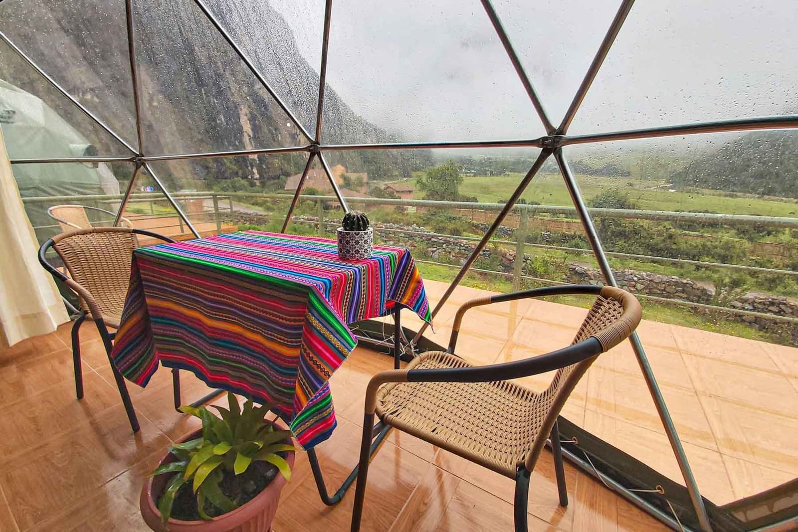 Chuva no domo, vale lá fora, e um cacto na mesa pra lembrar onde você está. Interior de um domo geodésico de madeira na trilha Salkantay de 5 dias, com mesa de jantar coberta por têxtil andino colorido, cadeiras de vime e painéis panorâmicos molhados de chuva com vista para um vale verde e montanhas rochosas
