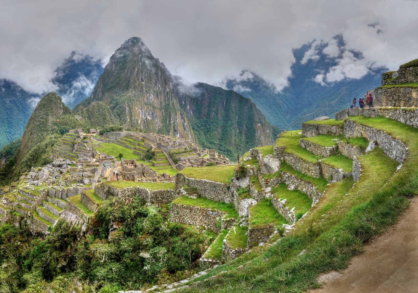 Seiscentos anos de pedra e as nuvens ainda não conseguiram cobrir tudo. Vista panorâmica da cidadela inca de Machu Picchu com terraços e ruínas de pedra espalhados por um cume, o pico Huayna Picchu emergindo por entre nuvens baixas ao fundo