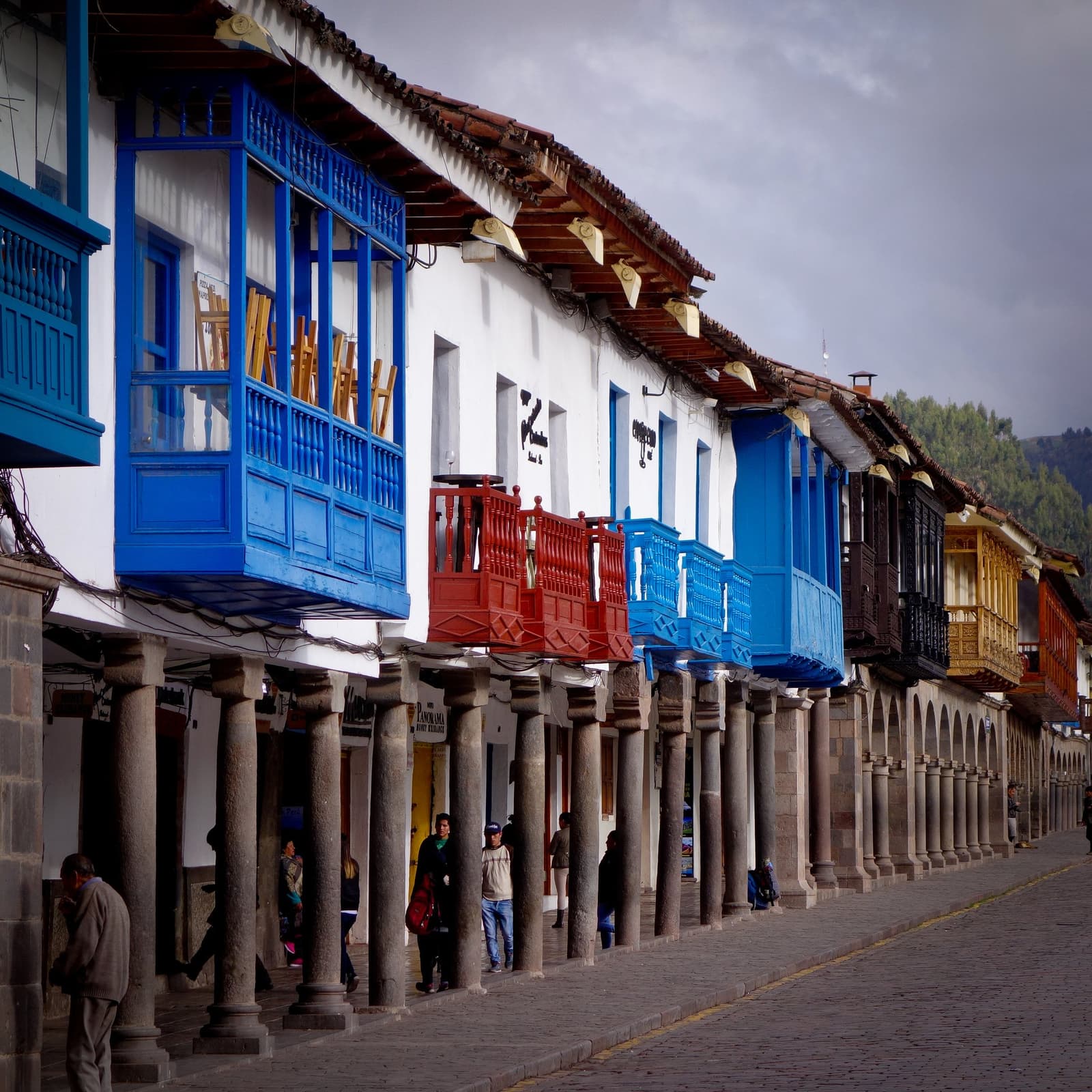 As varandas de Cusco já viram muitas chegadas. A sua é a próxima. Uma fileira de varandas de madeira coloridas em azul, vermelho e amarelo sobre arcadas de pedra em uma rua de paralelepípedos em Cusco