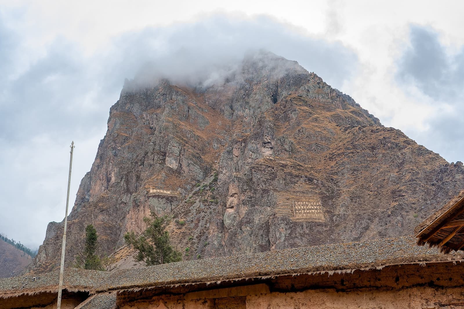 Celeiros cortados na rocha pura, porque os Incas nunca desperdiçaram uma parede. Photo: Brian Berg Armazéns e terraços incas esculpidos em um penhasco rochoso íngreme em Ollantaytambo, com nuvens baixas cobrindo o cume e telhados de adobe em primeiro plano