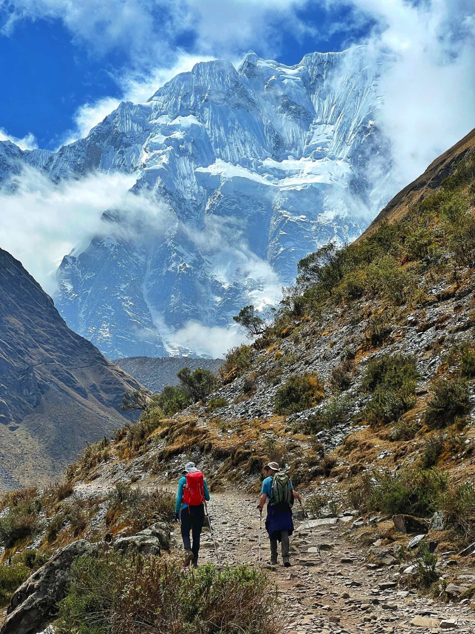 Dois caminhantes, uma montanha que ocupa todo o céu. É só continuar. Dois caminhantes com bastões e mochilas percorrem uma trilha rochosa na Salkantay trek para Machu Picchu, com um imenso pico nevado e glacial emergindo entre as nuvens à frente