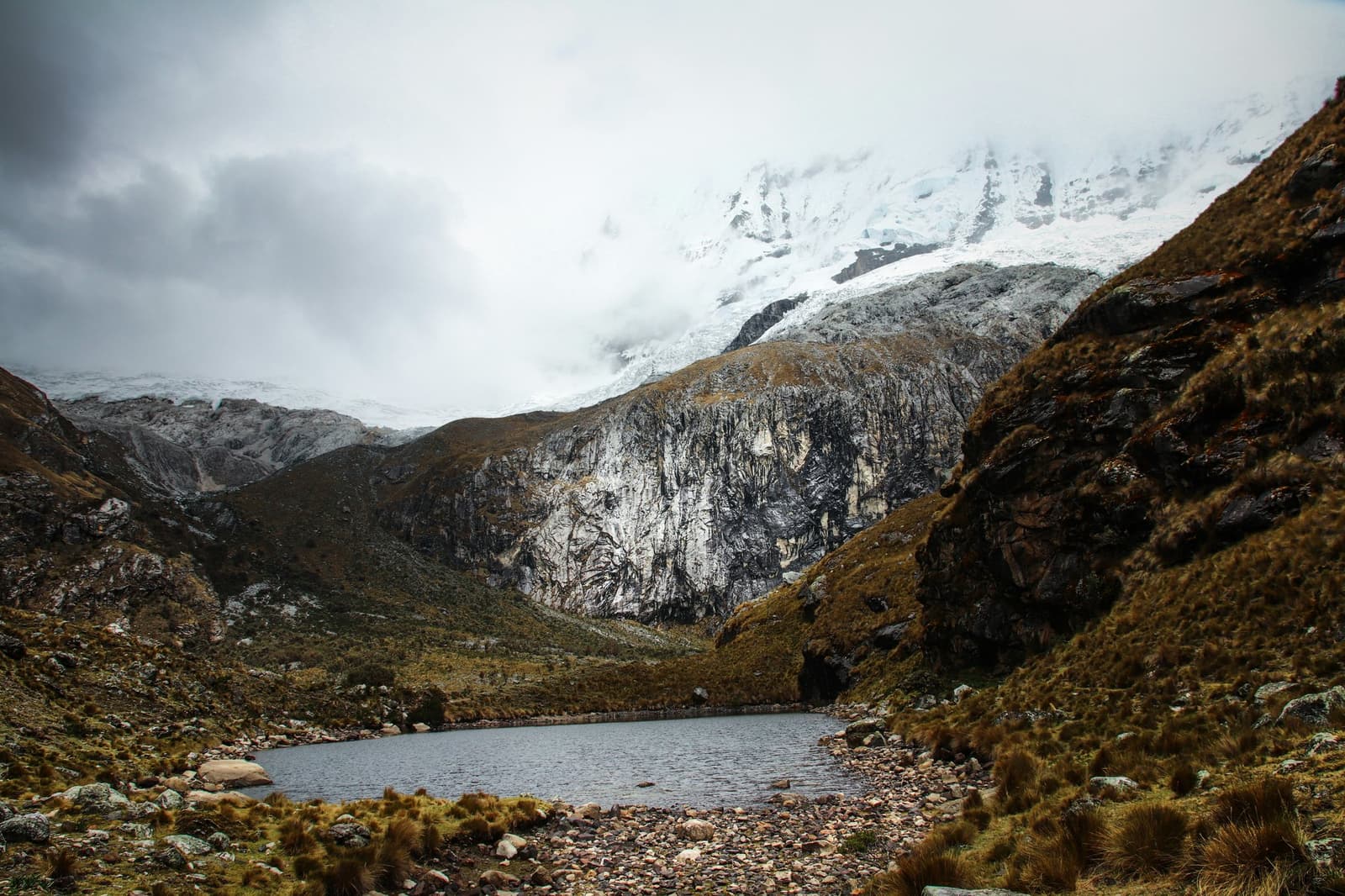 Frio, quieto e completamente alheio a quantos quilômetros você deixou para trás. Um pequeno lago glacial numa vale rochosa de alta altitude no trek de Santa Cruz, com picos nevados íngremes e nuvens baixas preenchendo o circo superior sobre encostas cobertas de gramíneas