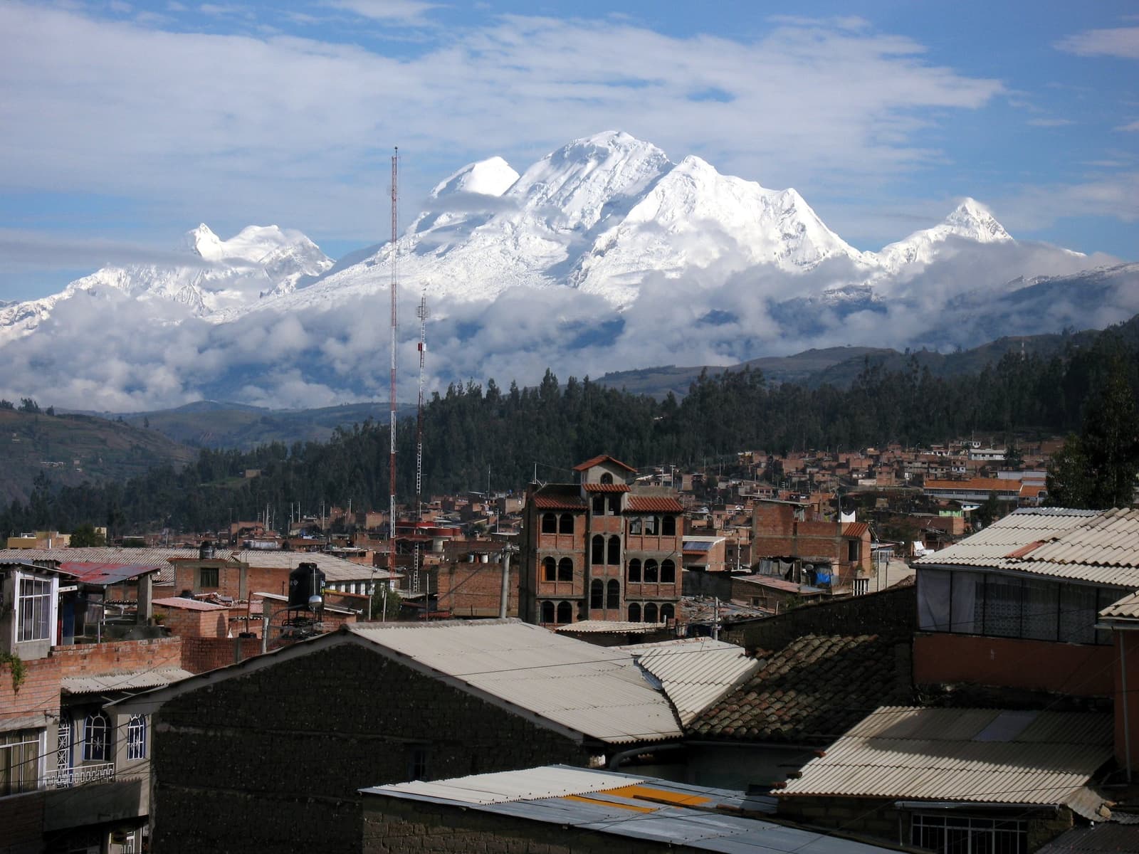 Um maciço glacial inteiro de pano de fundo, e a cidade segue sua rotina sem cerimônia. Photo: Uwe Behm Vista sobre os telhados de tijolos de uma cidade andina peruana com um grande maciço de montanhas nevadas acima da linha de árvores na rota do santa cruz trek saindo de Huaraz