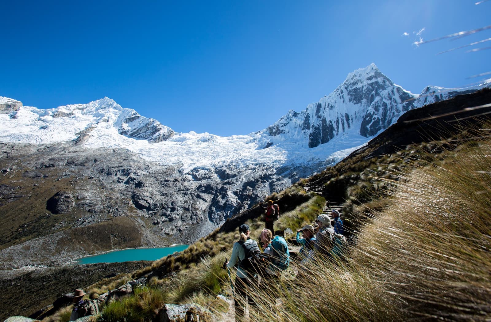 Legs earned that view. The glacial lake below is not going anywhere. A group of trekkers rests on a steep grass-covered trail during the santa cruz trek, with a vivid turquoise glacial lake and massive snow-capped peaks filling the valley below under a deep blue sky.